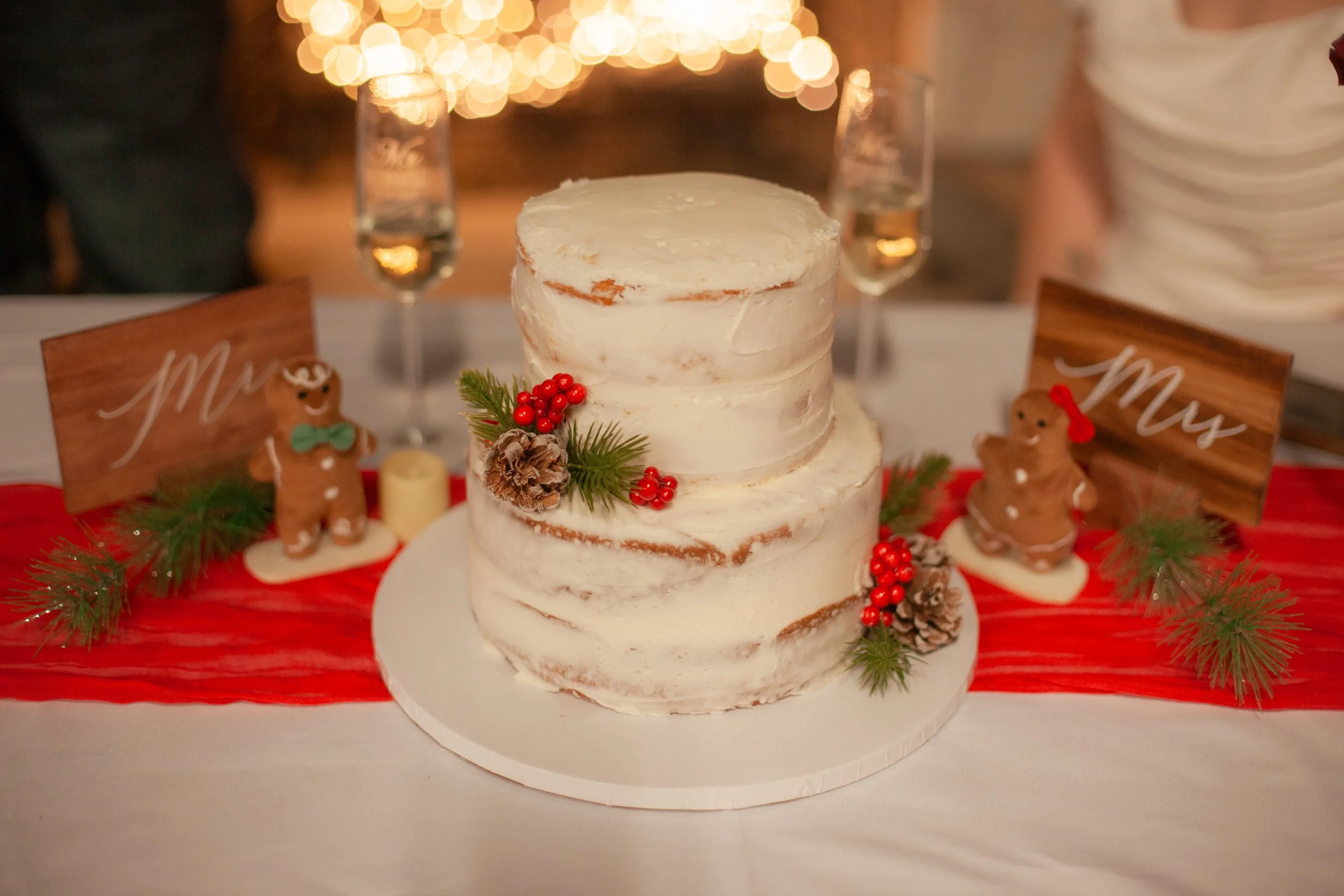 A two-tiered white wedding cake decorated with red berries, pinecones, and evergreen sprigs, set on a white cake board on a table with a red table runner, flanked by two wooden signs that read 'Mr' and 'Mrs' with gingerbread figures, and champagne gl