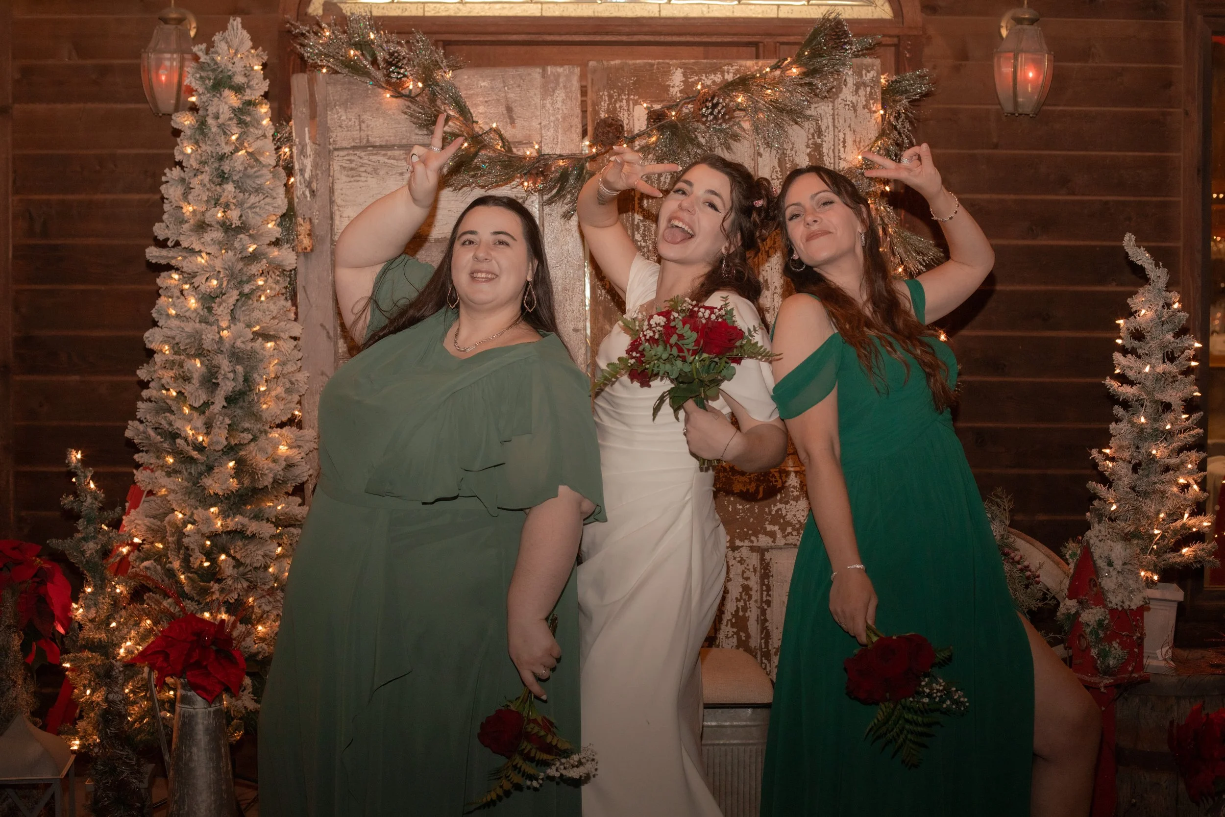 Three women at a wedding celebration, with one wearing a white dress and holding a bouquet of red roses, two in green dresses, celebrating in front of a decorated wooden backdrop with Christmas lights and pine garlands.