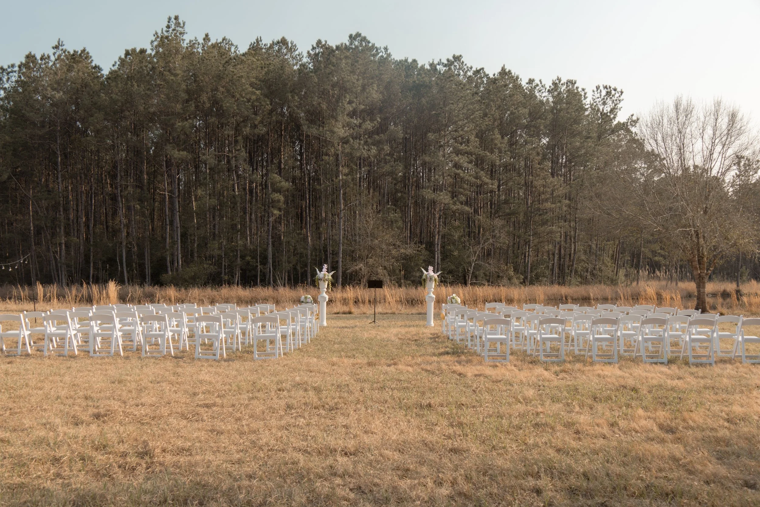 Outdoor wedding ceremony setup on a grassy field with white chairs arranged in rows on either side of an aisle, white decorative pillars with floral arrangements at the end of the aisle, set against a backdrop of trees and a partly sunny sky.