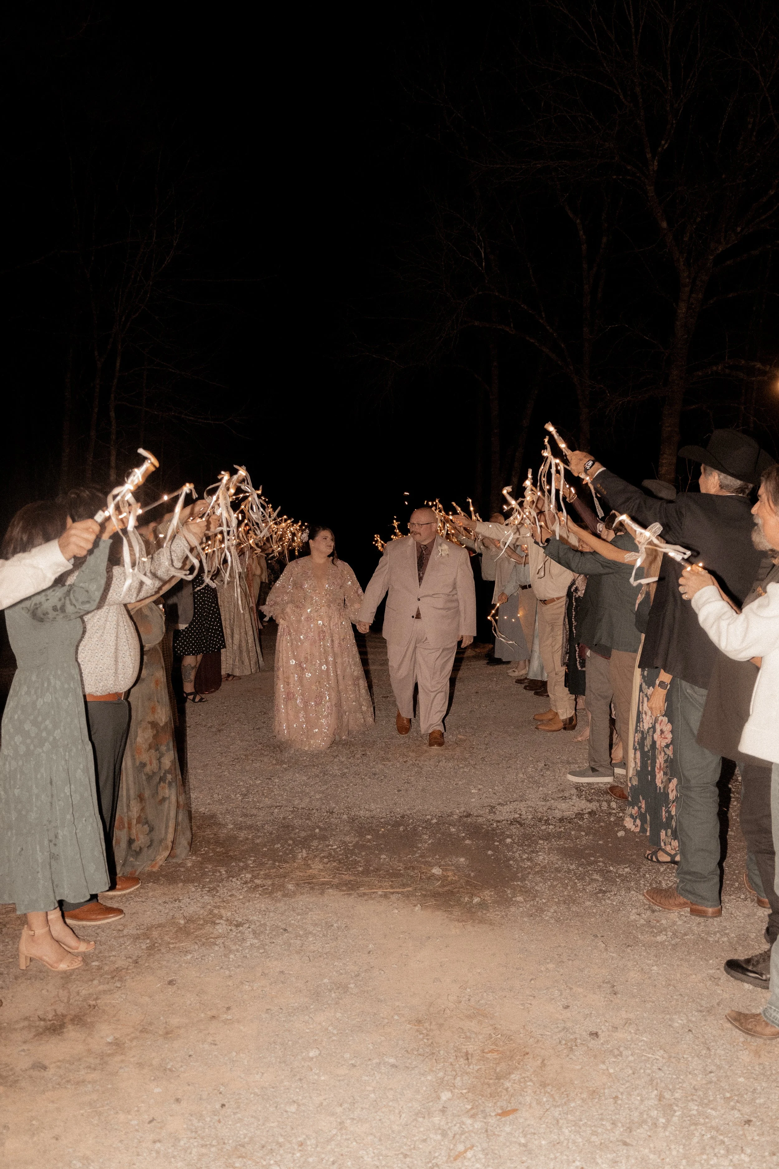 A couple walks hand in hand through a tunnel made of people holding sparklers at night, celebrating their wedding anniversary.