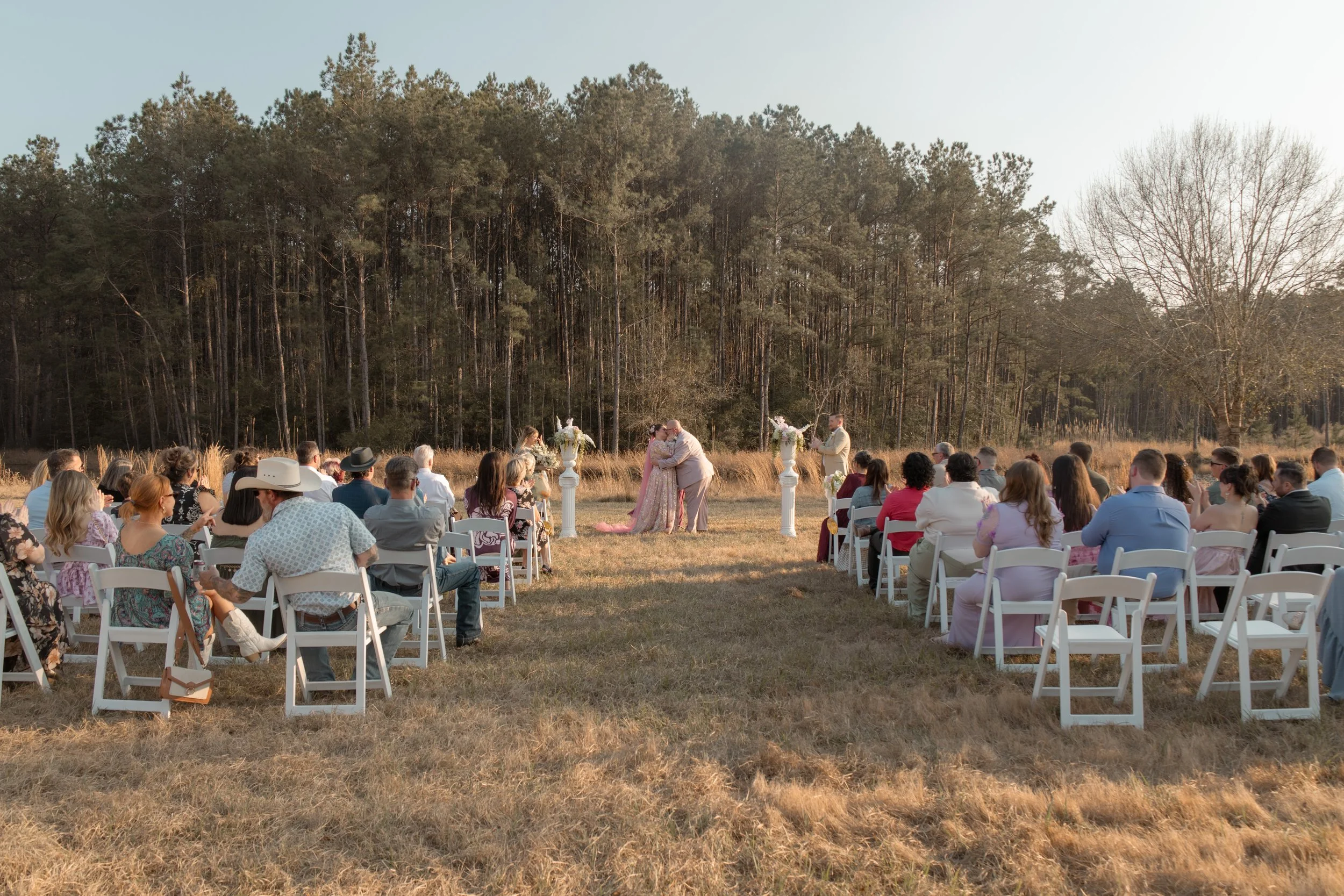 Outdoor wedding ceremony with guests seated on white chairs, a couple kissing at the altar, and two white pedestals with floral arrangements, set against a backdrop of trees and open sky.