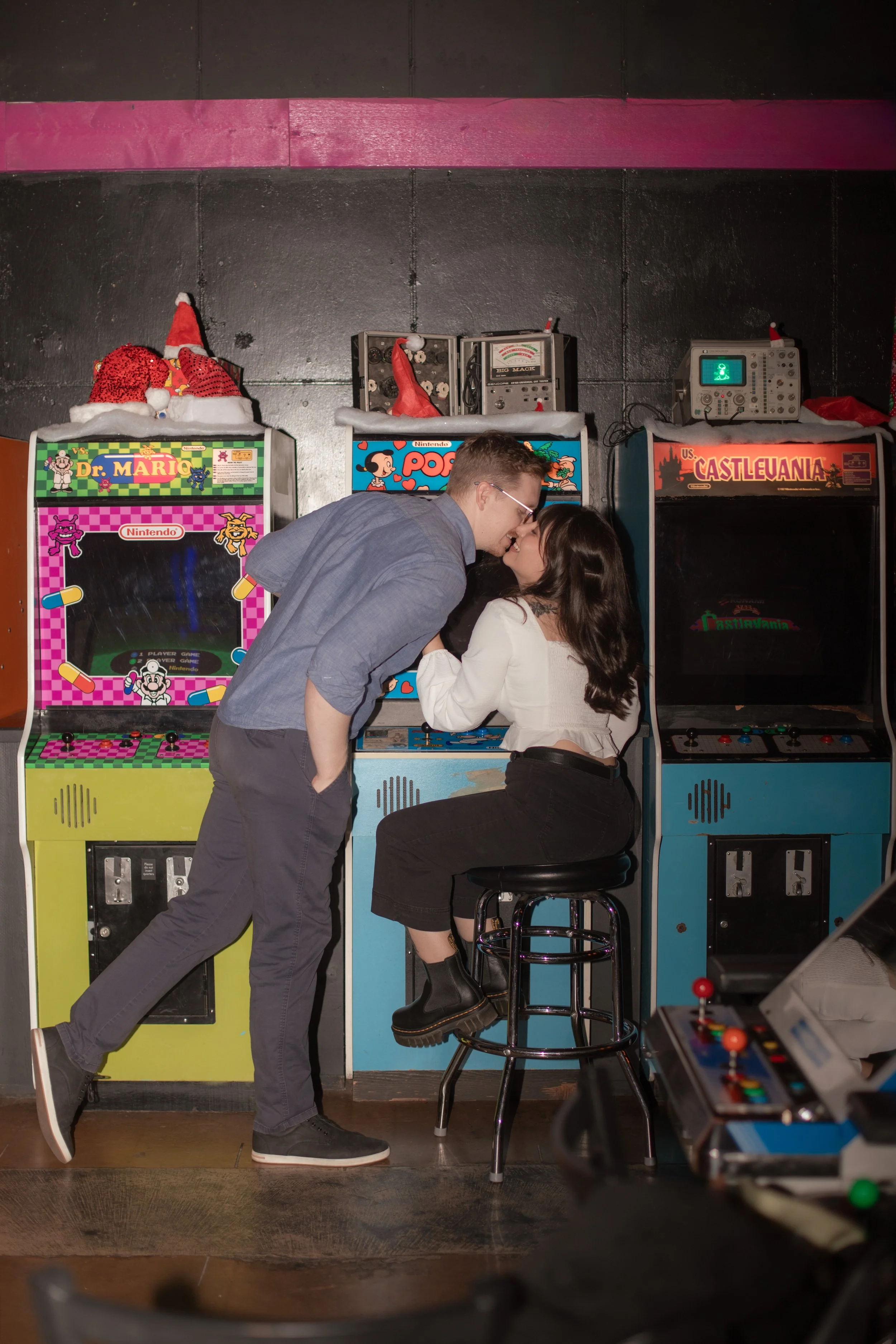 A couple sharing a kiss at an arcade, with vintage arcade machines visible behind them.