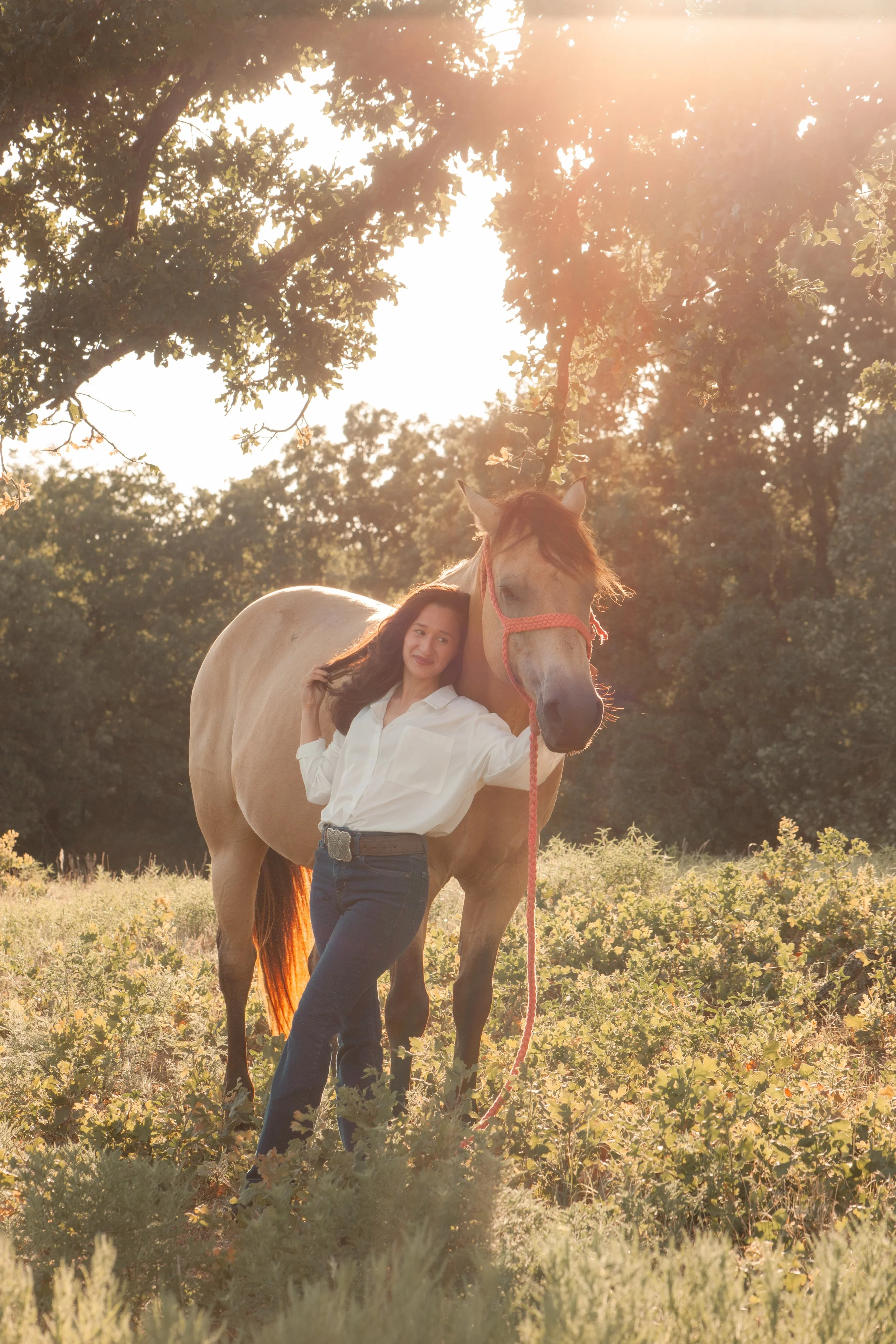 A woman standing next to a horse in a sunlit outdoor setting with trees and greenery.