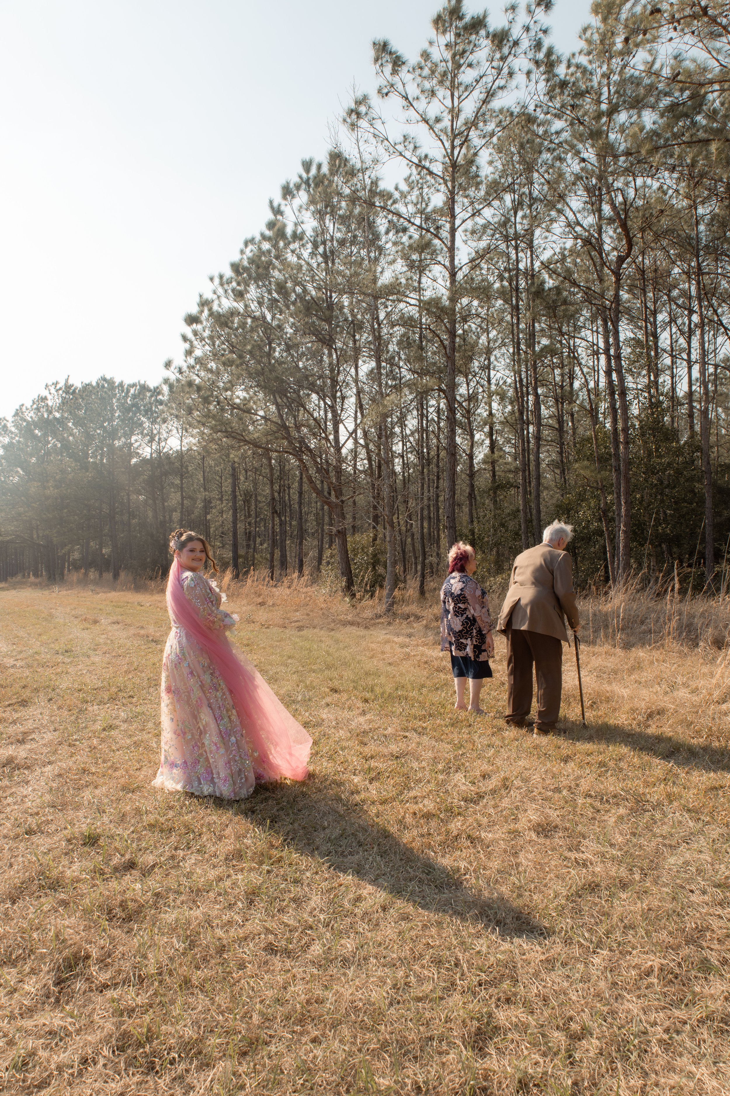A woman in a floral pink and cream dress with a pink veil walking on a grassy field, with two elderly people walking away from her toward a forested area, under tall trees on a sunny day.