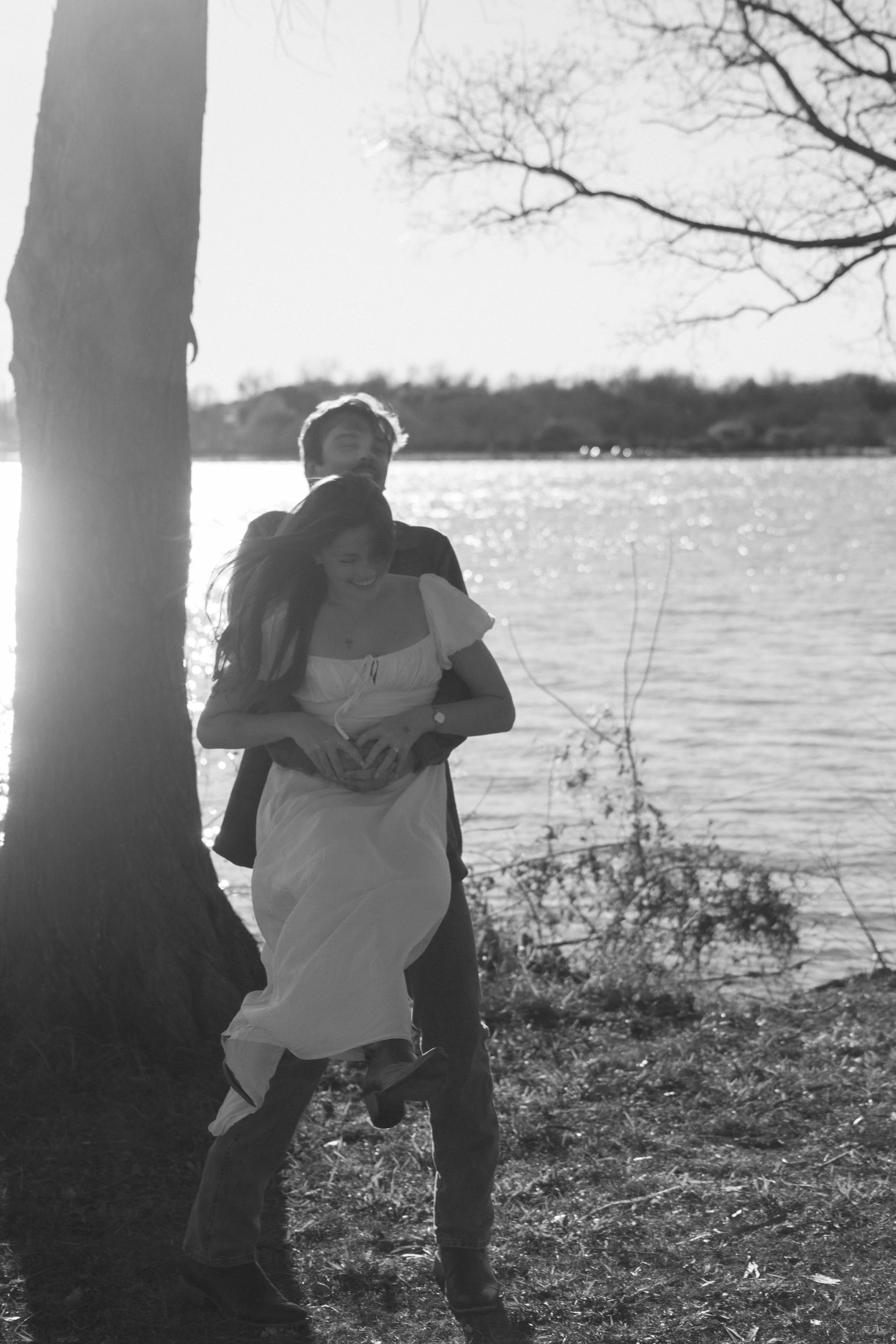 A couple standing near a tree by a lake during daylight, with the man lifting the woman and the woman smiling, both enjoying a moment together outdoors.
