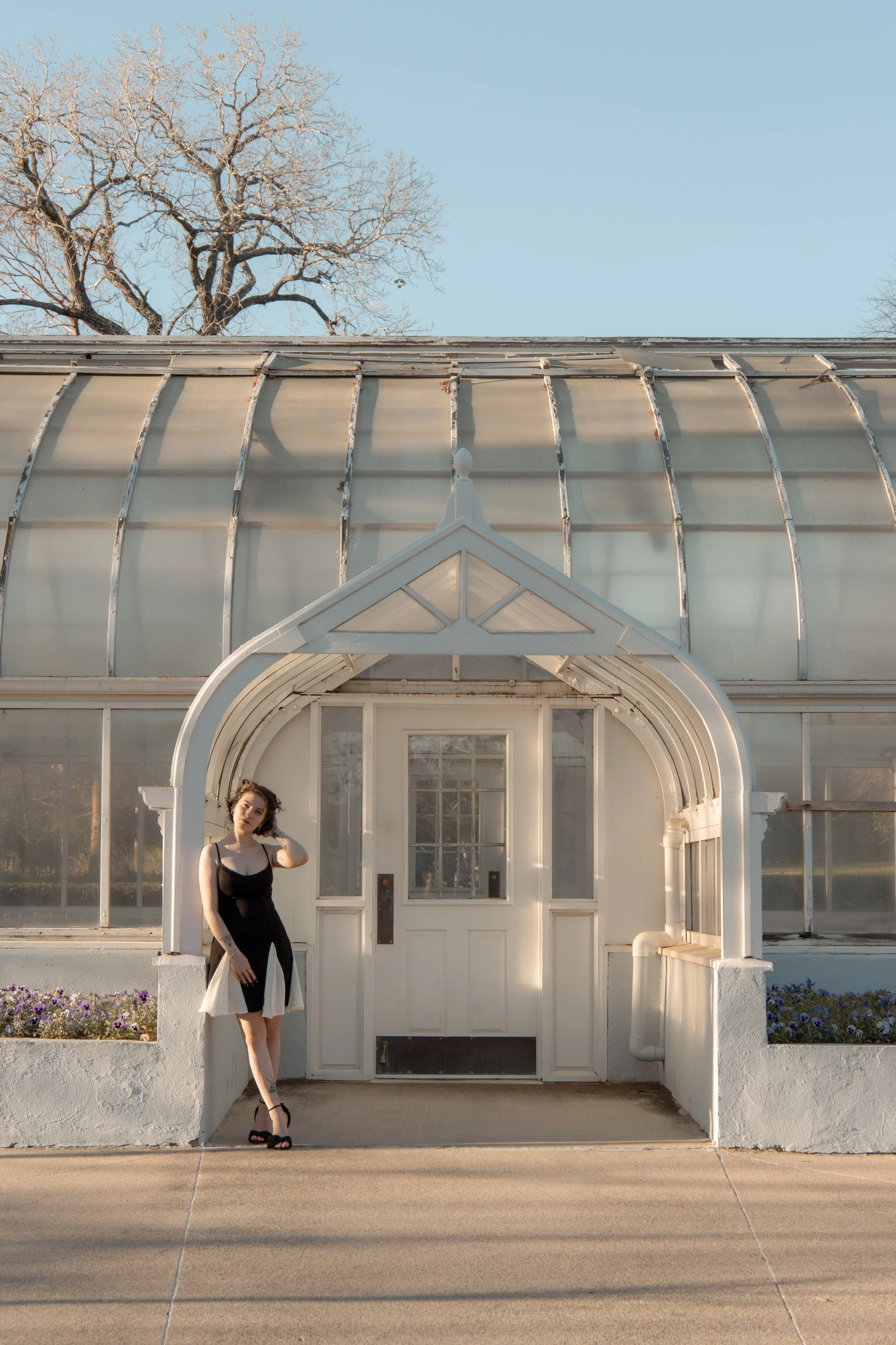 Woman in black and white dress standing outside a white greenhouse entrance during daytime.