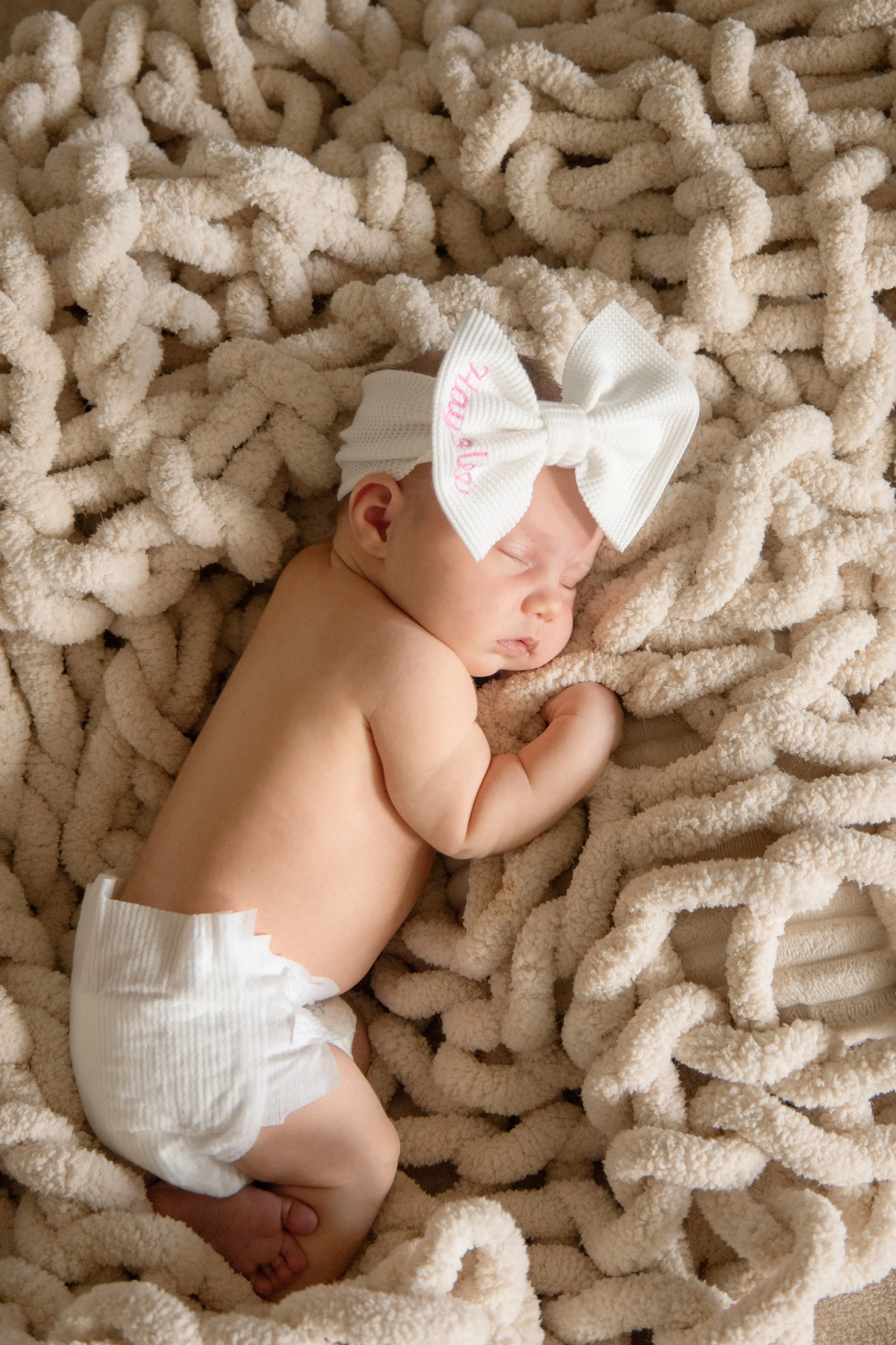 Newborn baby sleeping on a soft, beige blanket with a large white bow headband.