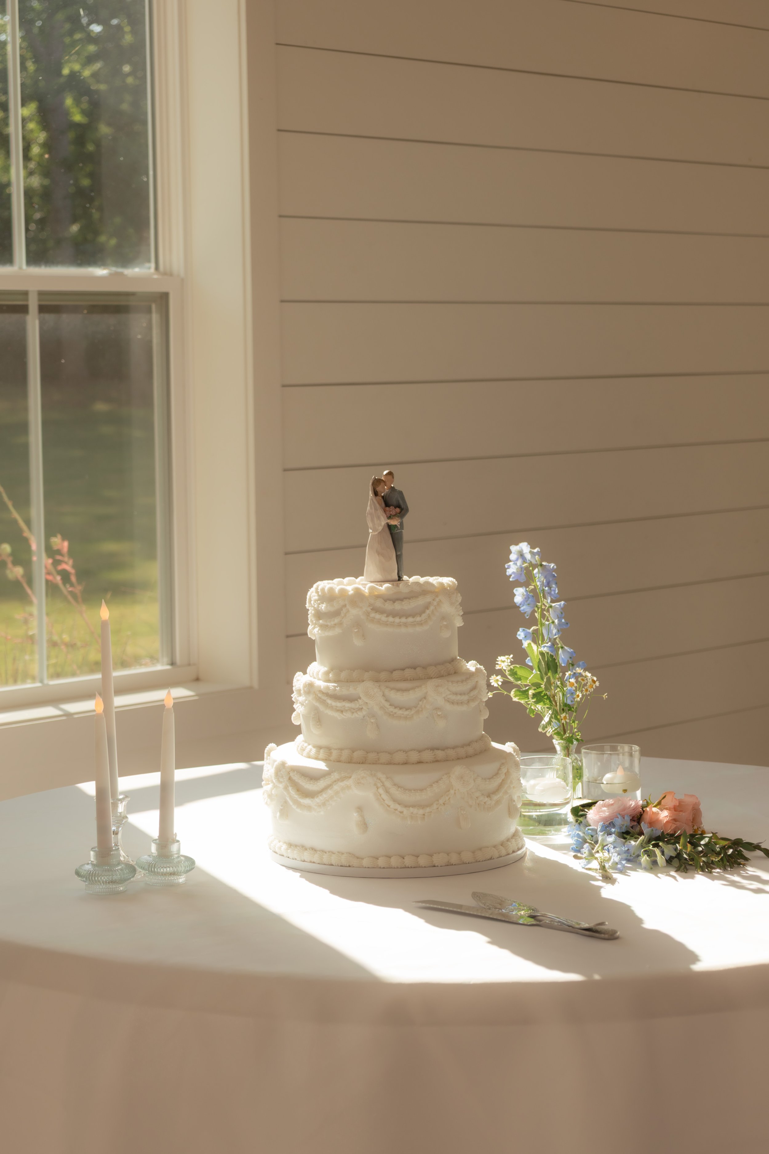 A multi-tiered white wedding cake with a bride and groom topper, surrounded by candles and flowers on a table near a window with sunlight.