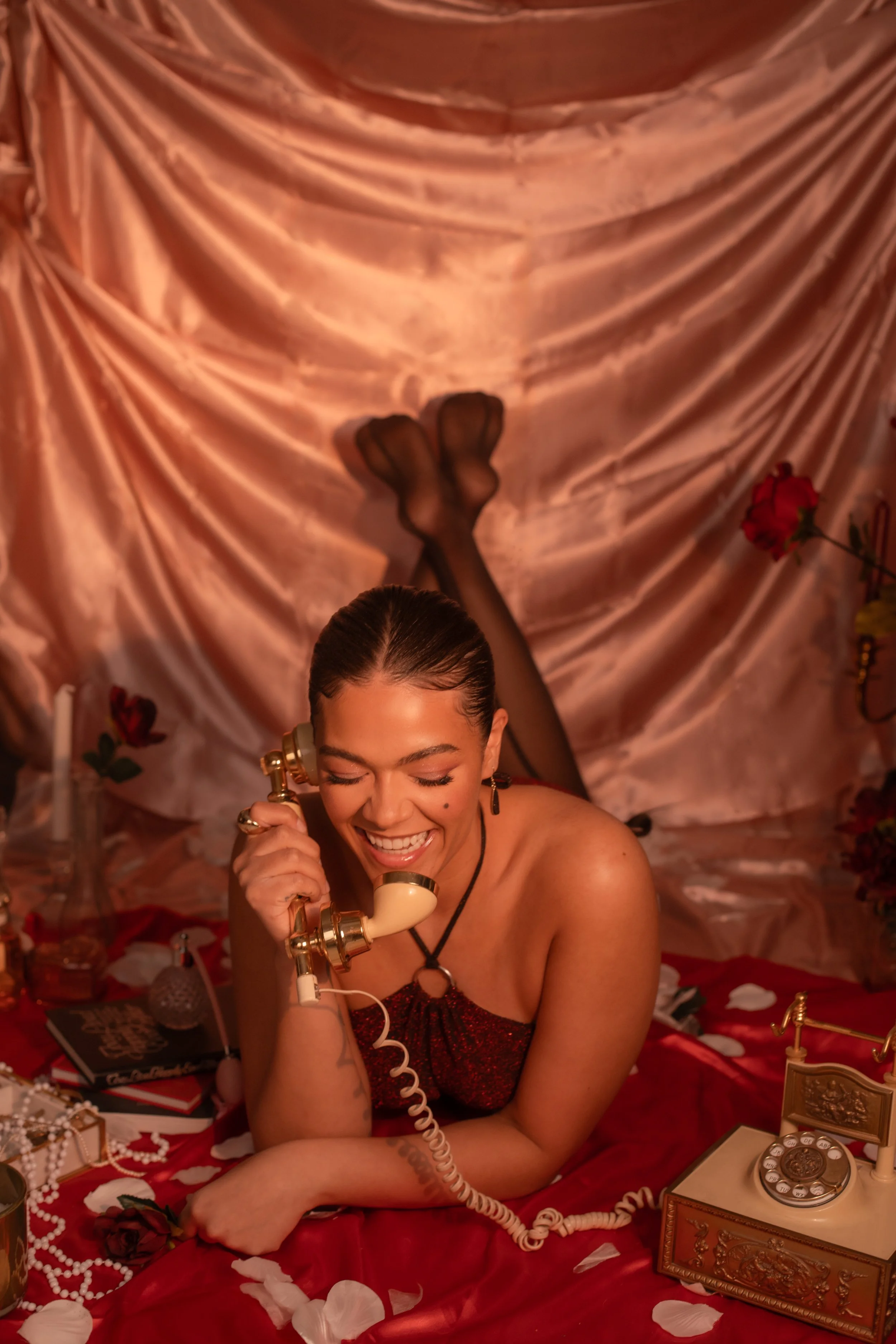 A woman lying on a bed, talking on a vintage rotary phone, smiling with eyes closed. She is surrounded by roses, pearls, and vintage items. The backdrop is silky pink fabric.