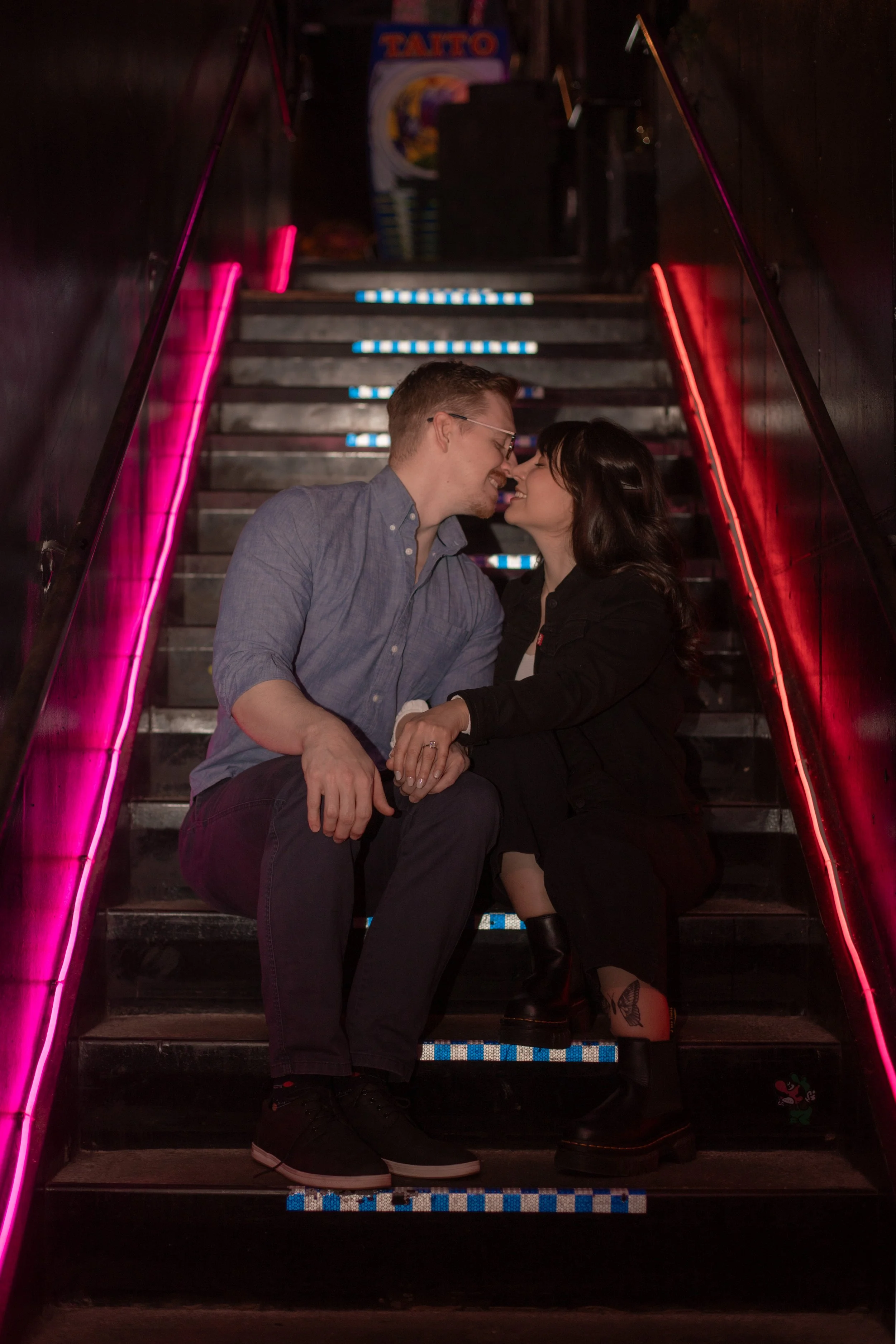 A couple is sitting on a staircase, nose to nose, laughing and holding hands, surrounded by pink and blue neon lights.