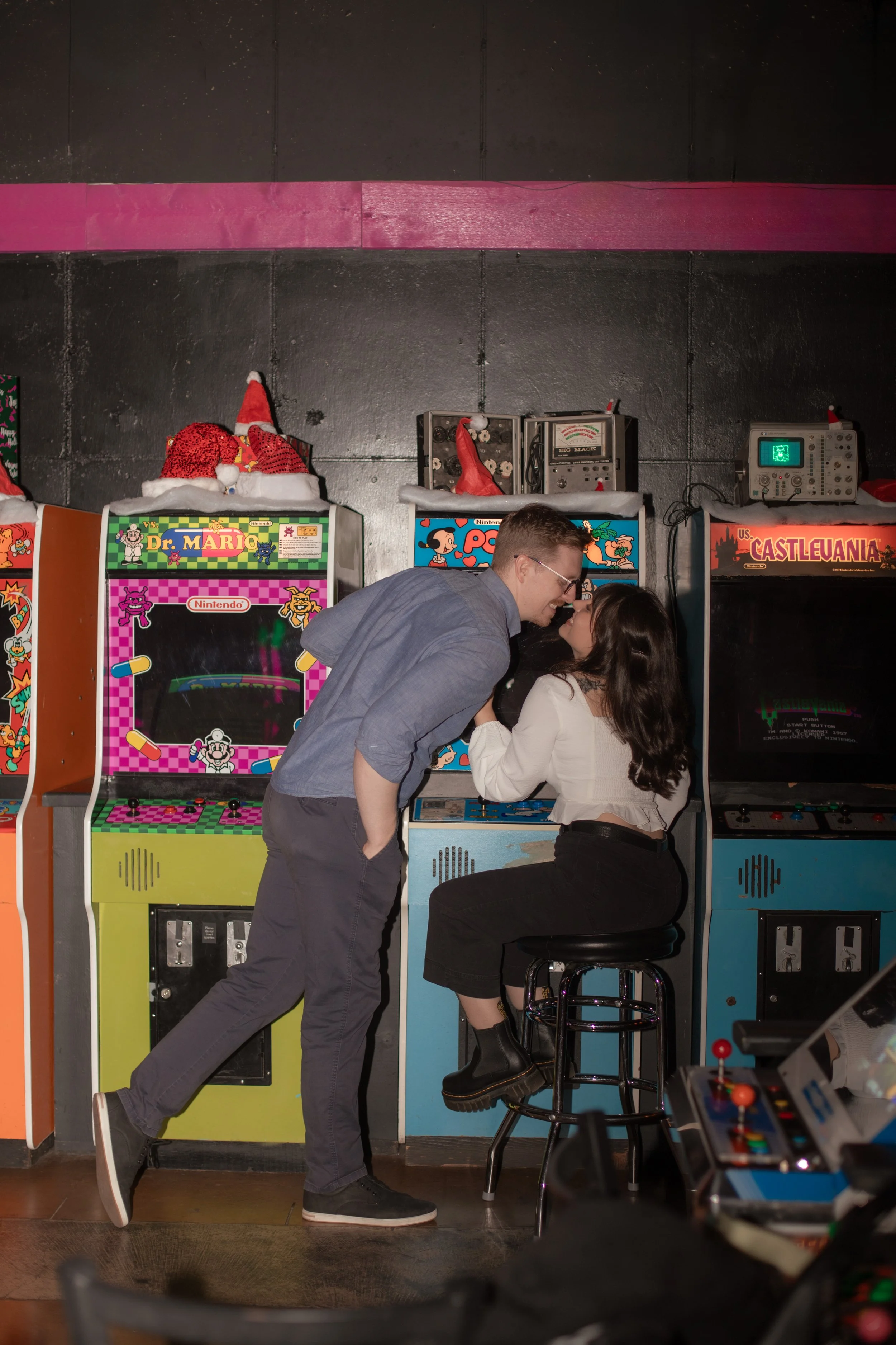 A couple sharing a kiss at an arcade with retro arcade game machines and Christmas Santa hats on top of the arcade cabinets.