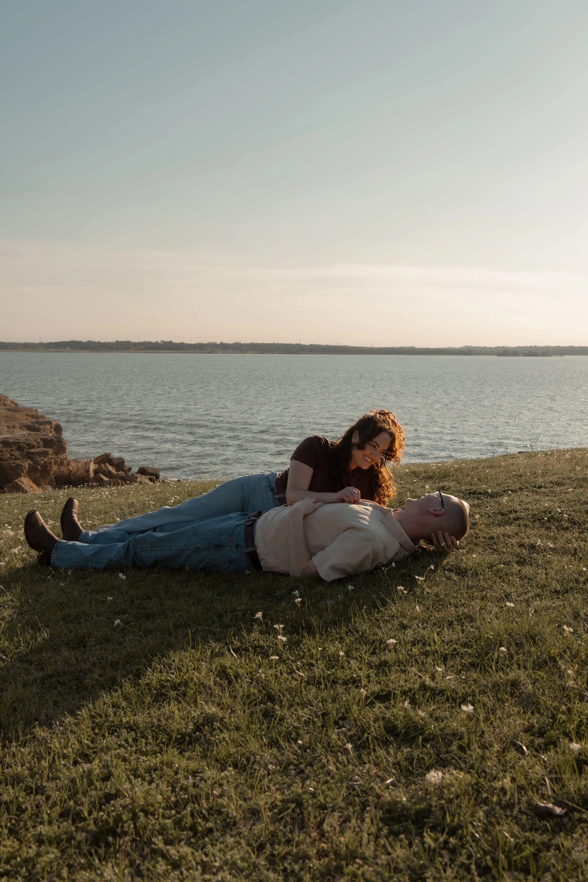 A woman and a man lying on grass near a body of water during sunset, the woman smiling and leaning on the man, who is lying on his back.