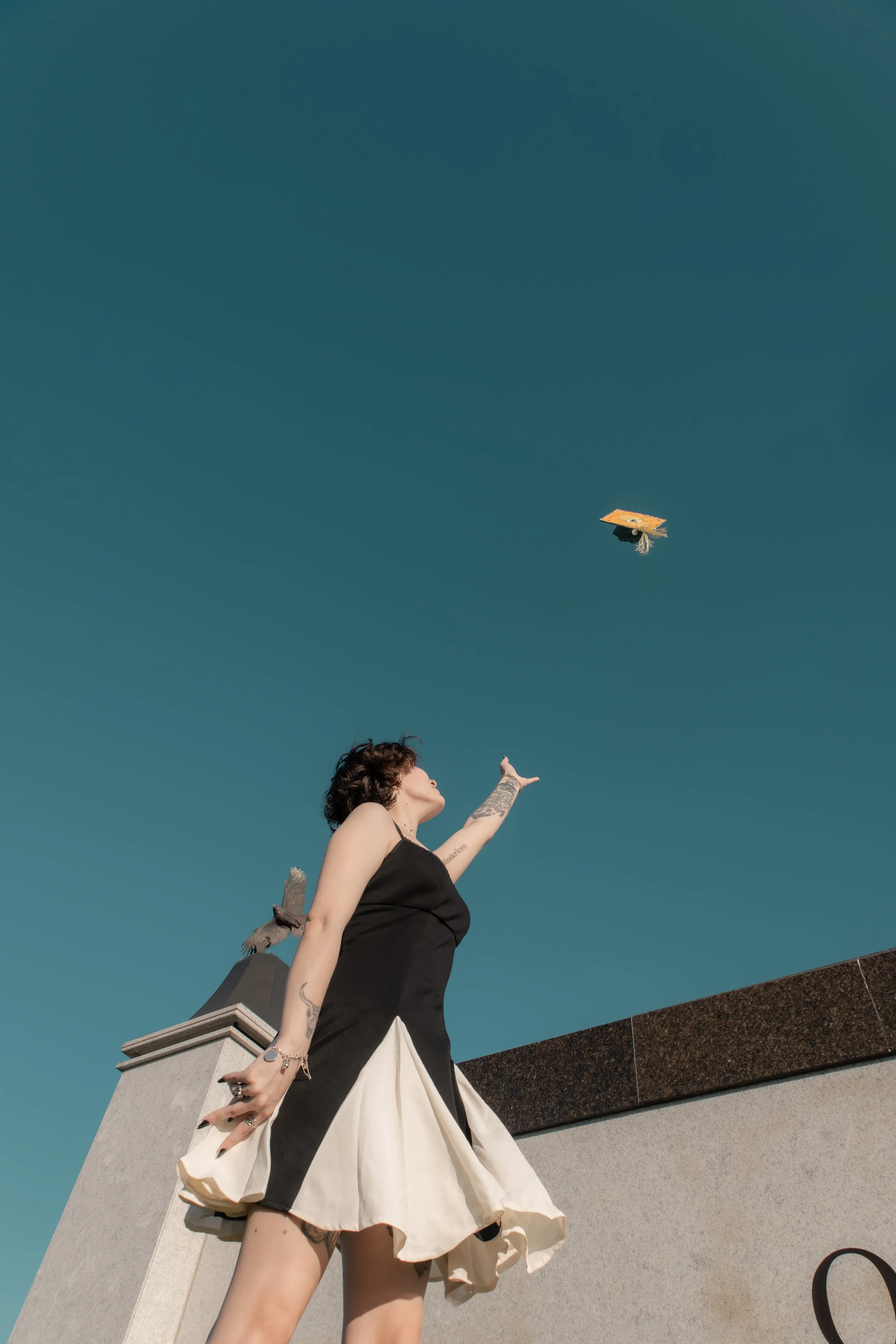A woman in a black and white dress throws a graduation cap into the air on a rooftop with a clear blue sky in the background.