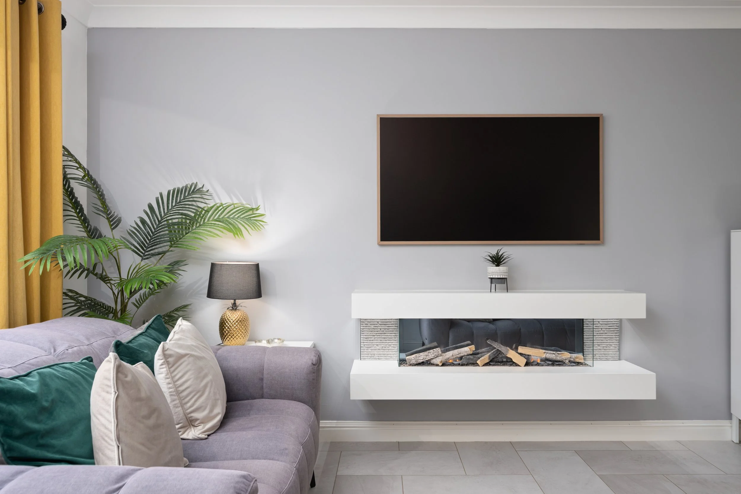 Living room with gray wall, modern fireplace with logs, large black-framed chalkboard, small plant on white mantel, gray sofa with green and white pillows, black table lamp with gold base, large green plant with palm leaves, yellow curtains, light-co