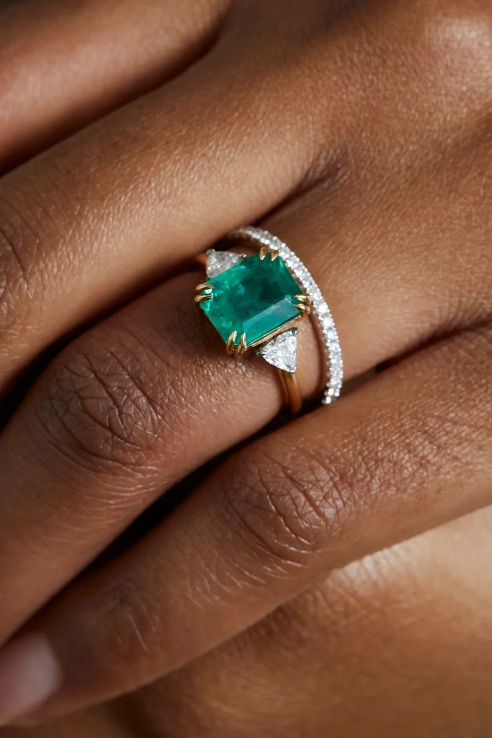 Close-up of a woman's hand with a large emerald ring, featuring a central emerald gemstone in a yellow gold setting, flanked by smaller diamonds, on her ring finger.