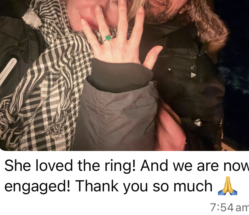 Close-up of a couple showing an engagement ring on the woman's finger, with the woman smiling and holding her hand up near her face, both wearing warm outdoor jackets.