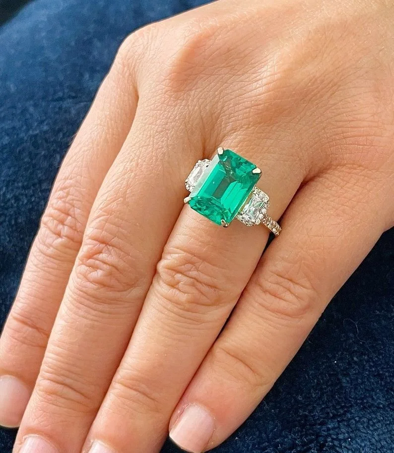Close-up of a hand wearing a ring with a large rectangular green gemstone flanked by two smaller clear gemstones on a silver band.