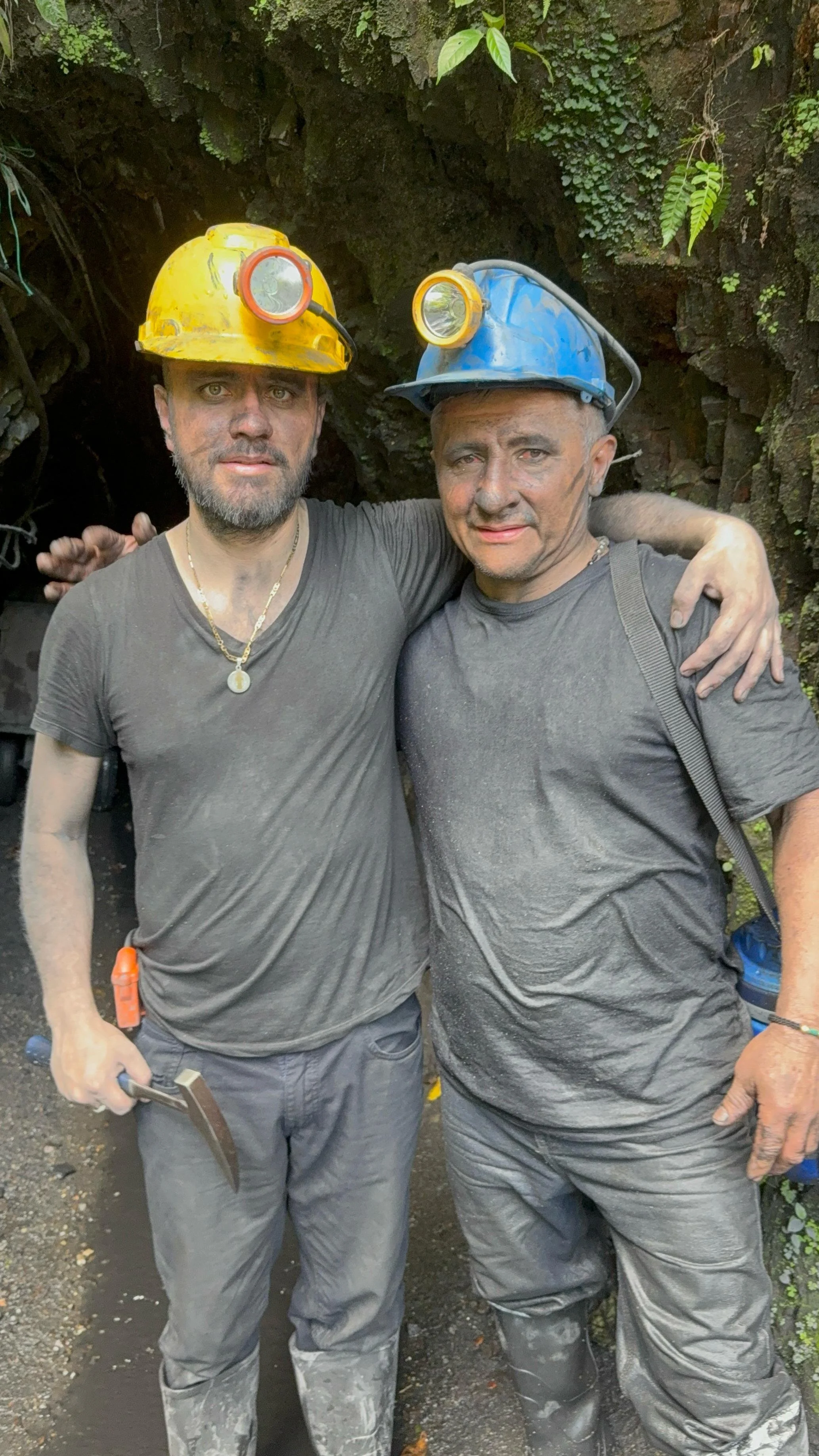Two miners wearing hard hats with lamps, standing close together inside a mine tunnel, and smiling at the camera.