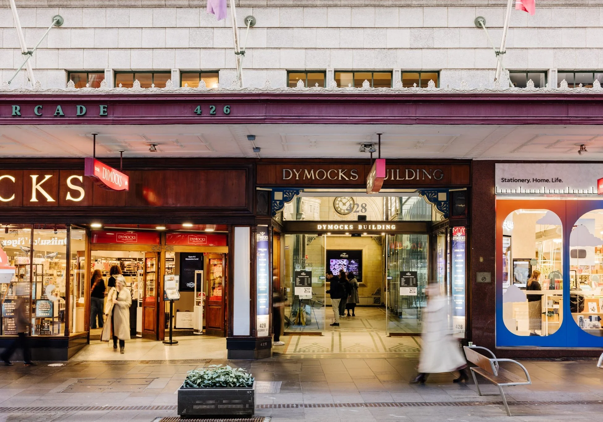 Entrance to Dymocks bookstore building located at 426, with people walking by on the sidewalk and storefront windows displaying books and stationery. Signage includes the store name and office building name.
