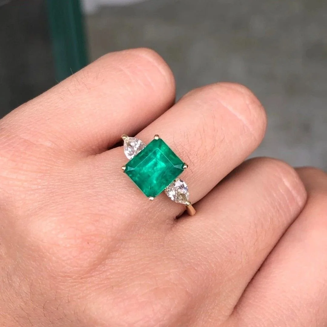 A gold ring with a large, cushion-cut green emerald gemstone in the center, flanked by two smaller pear-shaped diamonds on each side, being worn on a person's finger.