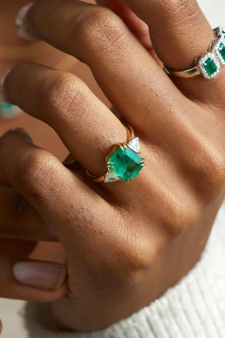 Close-up of a person's hand with rings, including a large green gemstone ring and a silver ring with green stones.