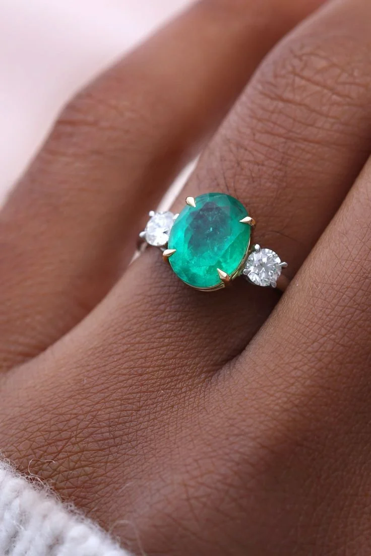 Close-up of a ring with an oval green gemstone and two smaller clear diamonds on either side, worn on a person's finger.
