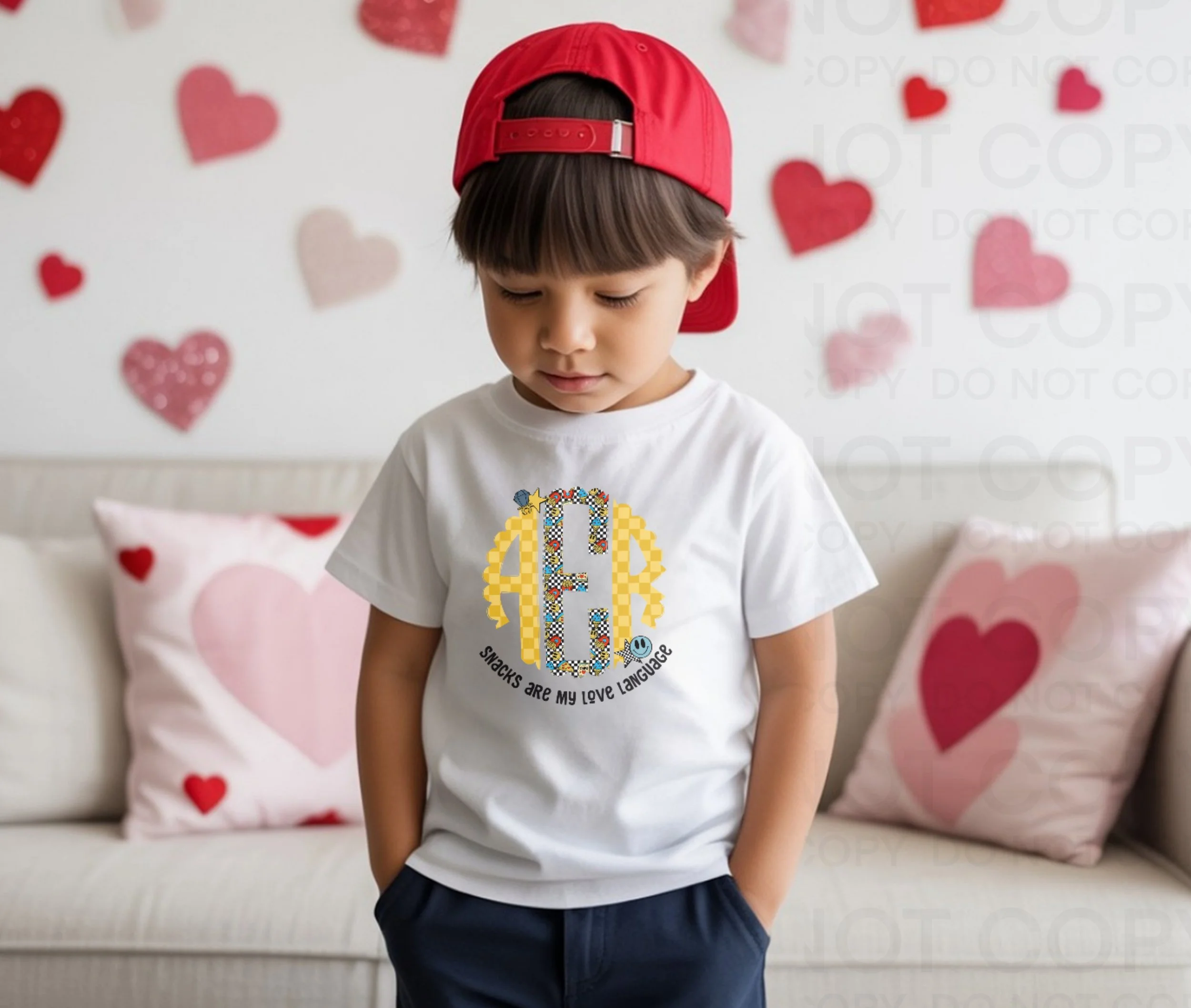 A young boy wearing a white T-shirt with a letter 'C' and the phrase 'Snacks are my love language,' standing in front of a sofa with pillows adorned with hearts. He is wearing a red backward baseball cap.