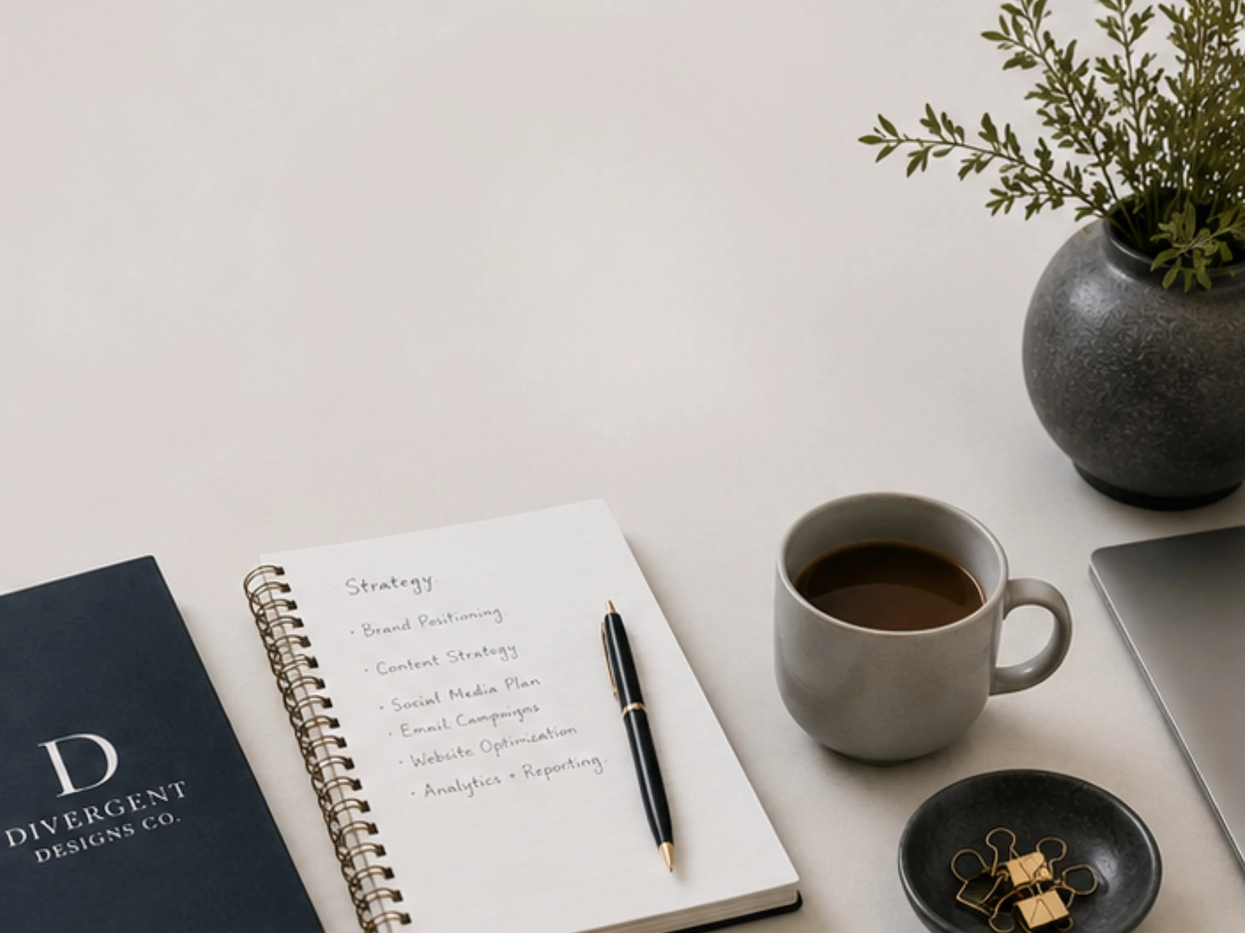 A desk workspace with a spiral-bound notebook labeled 'Strategy' open on the page listing marketing topics, a pen resting on the notebook, a cup of coffee, a large stone-gray vase with greenery, a black binder with white text, and a smartphone placed nearby.