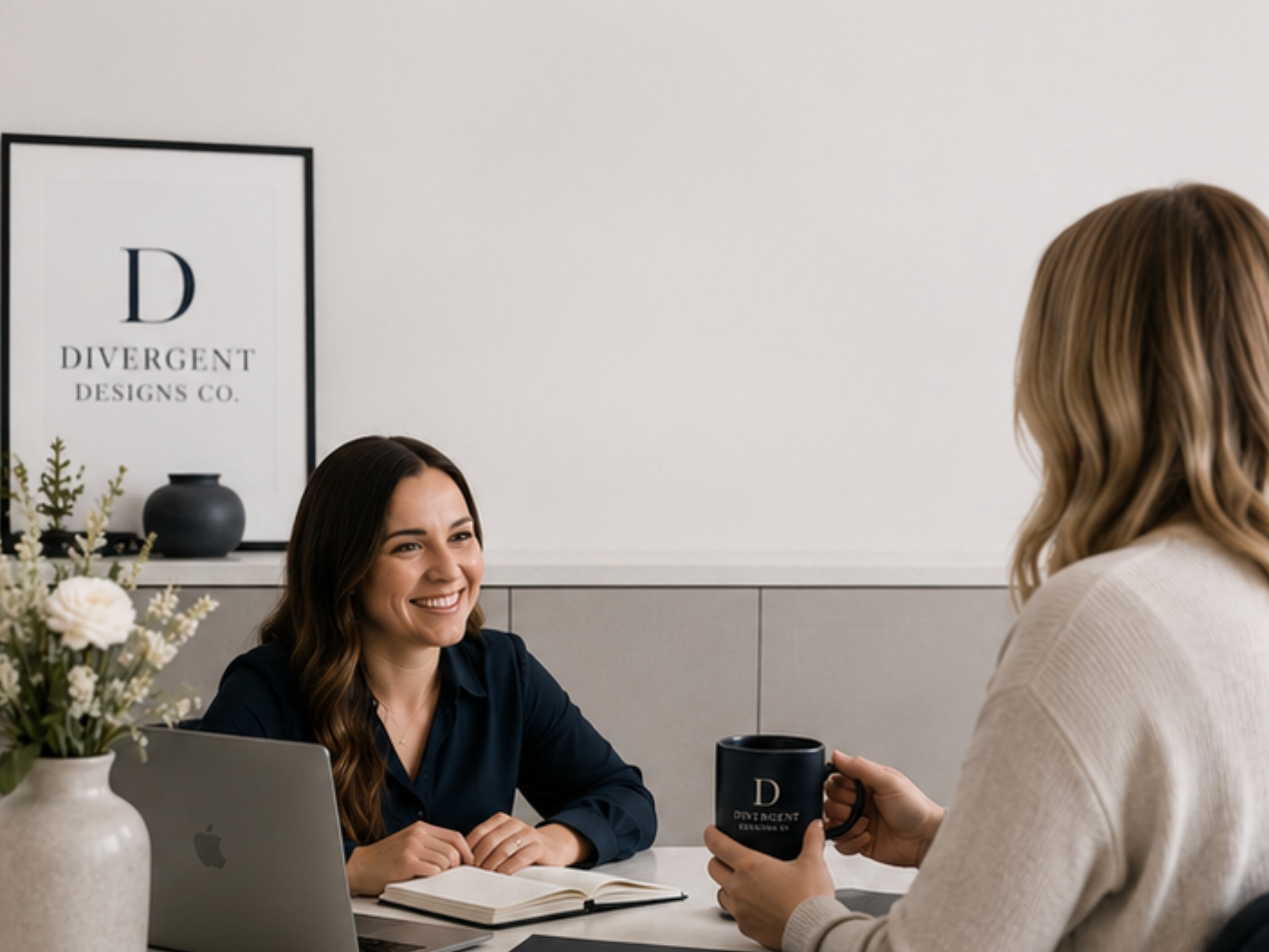 A woman with long dark hair smiling while sitting at a table, receiving a mug that says 'Divergent Designs Co.' from another woman with wavy blonde hair. On the table are a laptop, an open notebook, a black mug, and a white vase with flowers. A framed sign on the wall reads 'Divergent Designs Co.'.