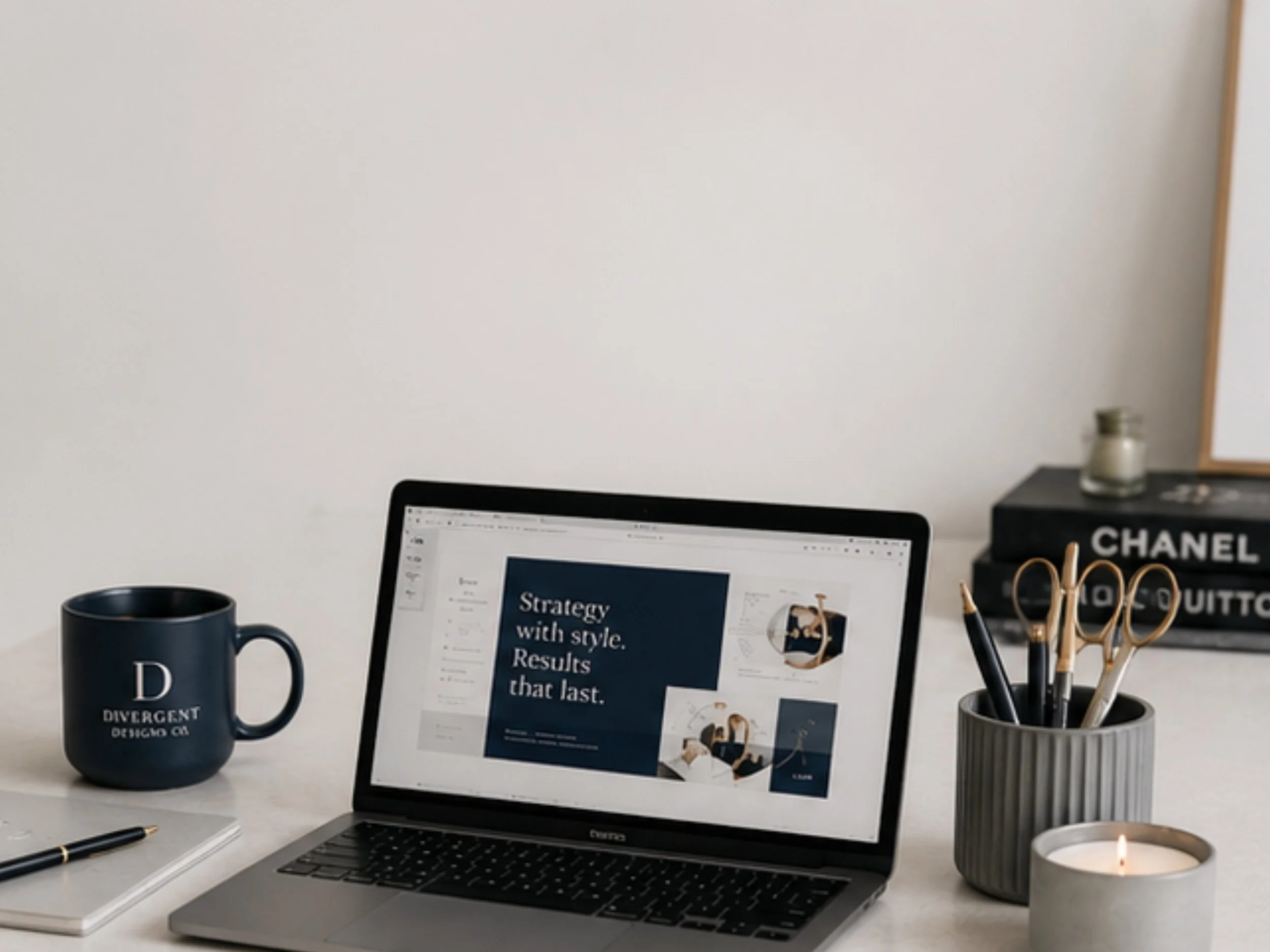 A workspace with a black laptop displaying an open presentation, a blue coffee mug with white text, a notepad with a black pen, a black organizer holding gold pens, a candle, and a black box with white text, all arranged on a white desk against a plain wall.