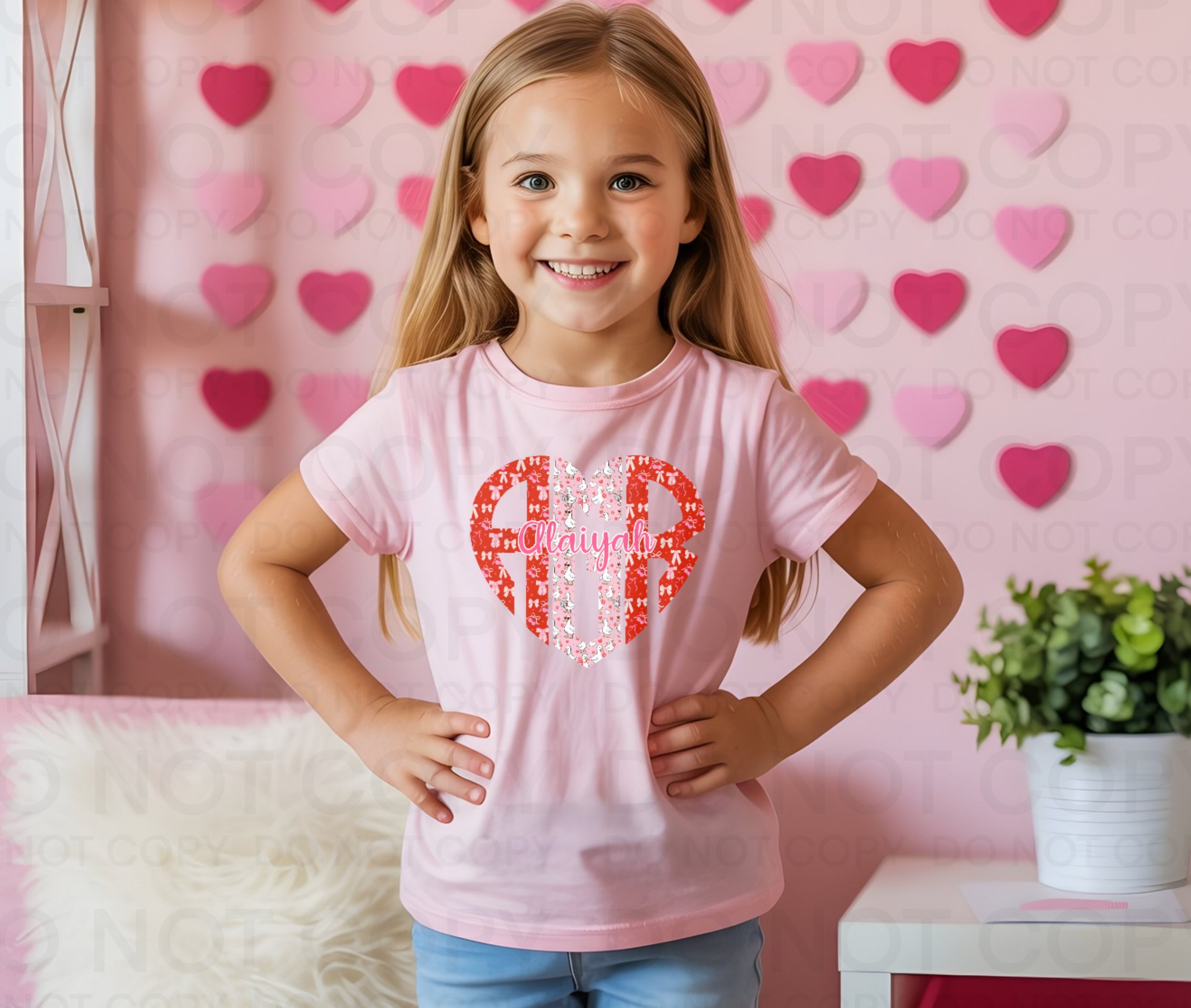 A smiling young girl with long blonde hair, wearing a pink T-shirt with a red and white heart design and the name 'Aaliyah' inside, stands with her hands on her hips in front of a pink wall decorated with paper hearts. There is a white table with a potted plant on it to her right.