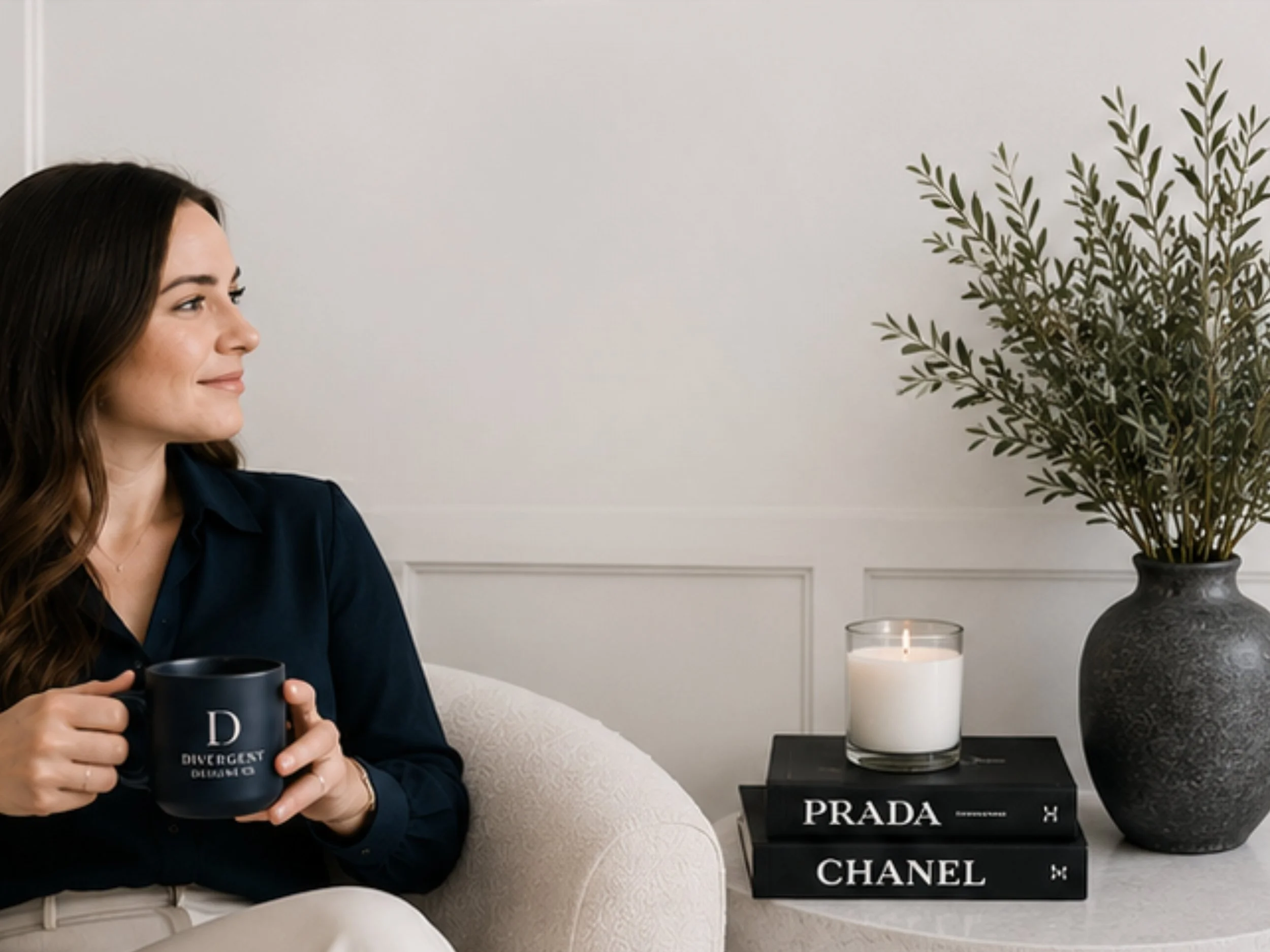 A woman sitting on a cream sofa holds a black mug with white text. Next to her is a side table with two stacked fashion books, a lit candle, and a tall gray vase with green leafy branches, all against a white wall with decorative paneling.