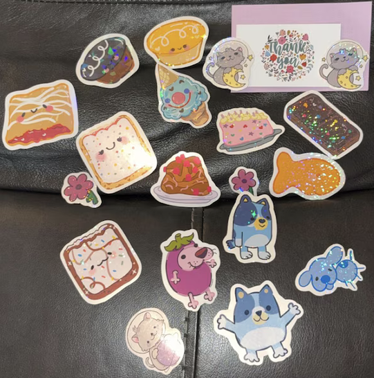 A collection of colorful stickers featuring animals, desserts, flowers, and a thank you card on a black surface.