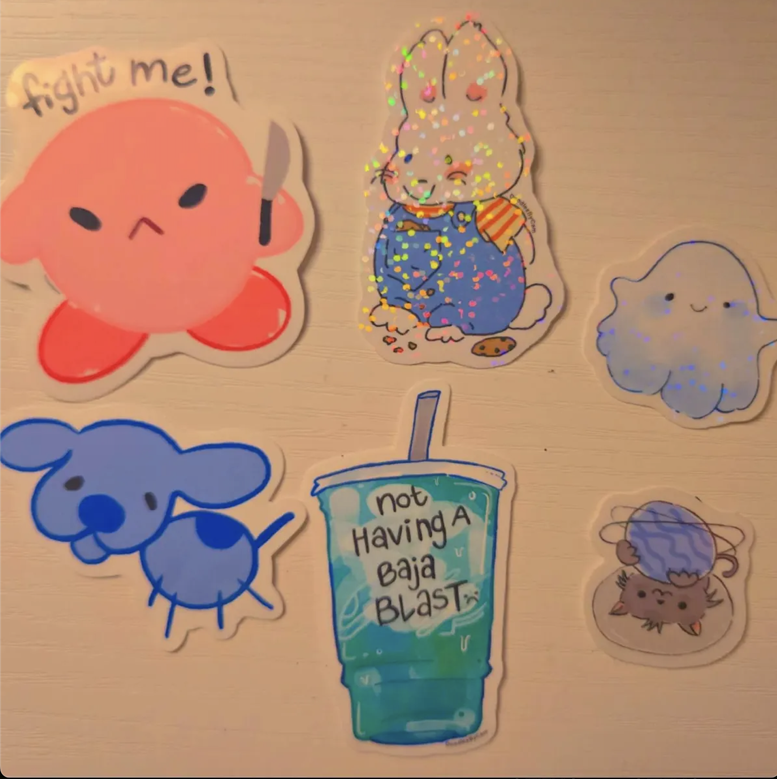 Collection of colorful cartoon stickers on a light-colored surface, featuring characters with expressive faces, a drink with the message "not Having A Baja BLAST," and various animated creatures.