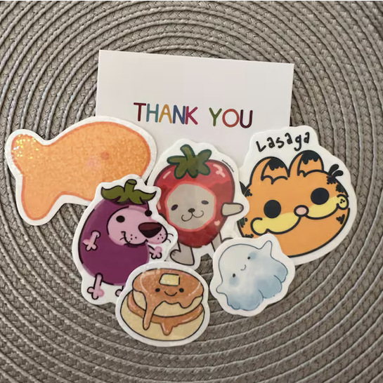 Collection of colorful stickers, including a fish, purple dog, strawberry, orange cat, lasagna, pancake, and a blue fluffy object, with a thank you card in the background.