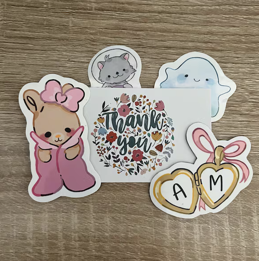 Collection of stickers including a cute bear with a pink bow, a gray cat, a smiling blue jellyfish, a thank you card with floral design, and two heart-shaped tags with initials A and M.