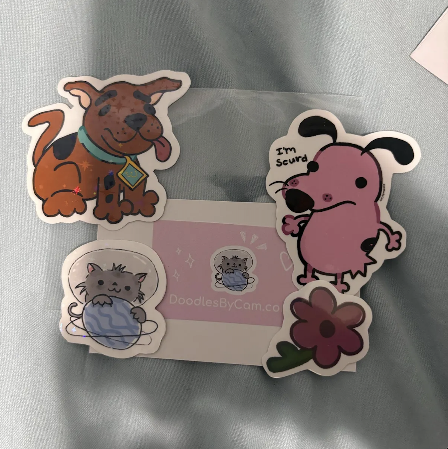 Collection of four cartoon stickers on a silver surface, featuring a brown dog with a green collar, a pink dog with black ears and a speech bubble saying 'I'm scared,' a grey cat holding a ball of yarn, and a large pink flower with green leaves.