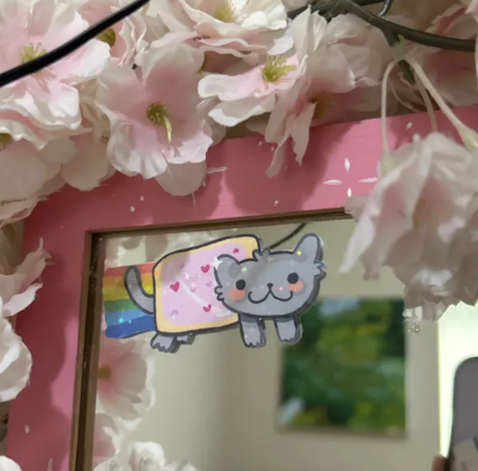 Cute sticker of a gray cat with pink cheeks and a happy face, connected to a rainbow, placed on a pink frame surrounded by light pink cherry blossoms.