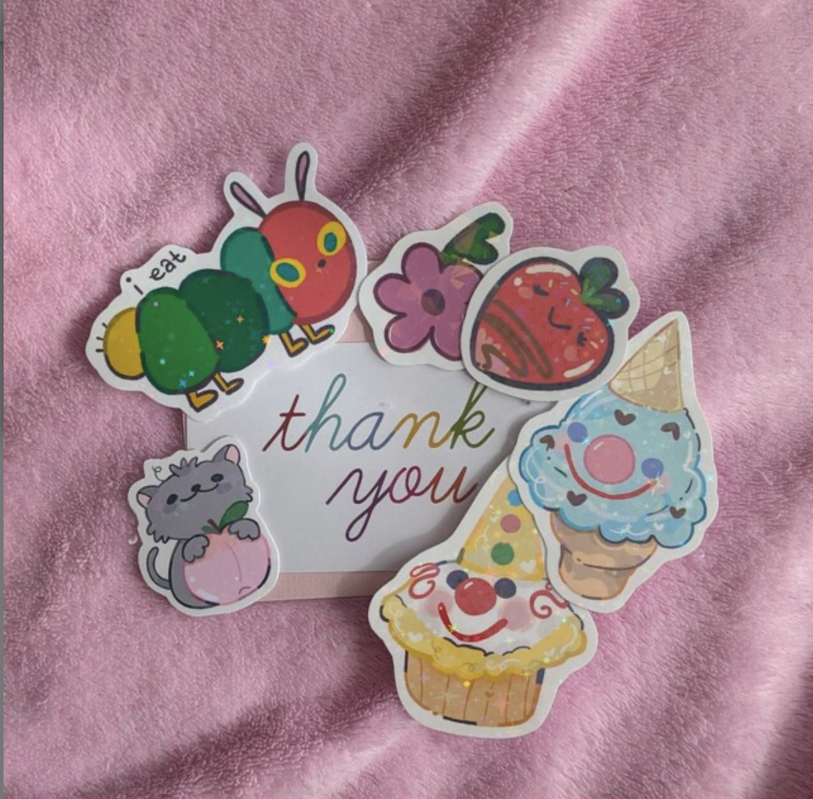Colorful thank you card with various cartoon stickers, including a strawberry, an ice cream cone with a clown face, a purple flower, a cute gray cat holding a peach, a red and green cartoon insect, and a cute red squiggly character, all on a pink textured fabric background.