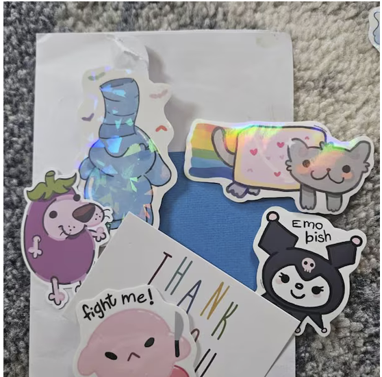A collection of colorful stickers on a blue folder or paper. The stickers include a rainbow-colored unicorn, a smiling gray cat with pink cheeks, a purple dog with a green leaf on its head, a black and white creature with horns and the text "emo bish," and a pink character with the text "fight me!" along with a handwritten note that says "THANK".