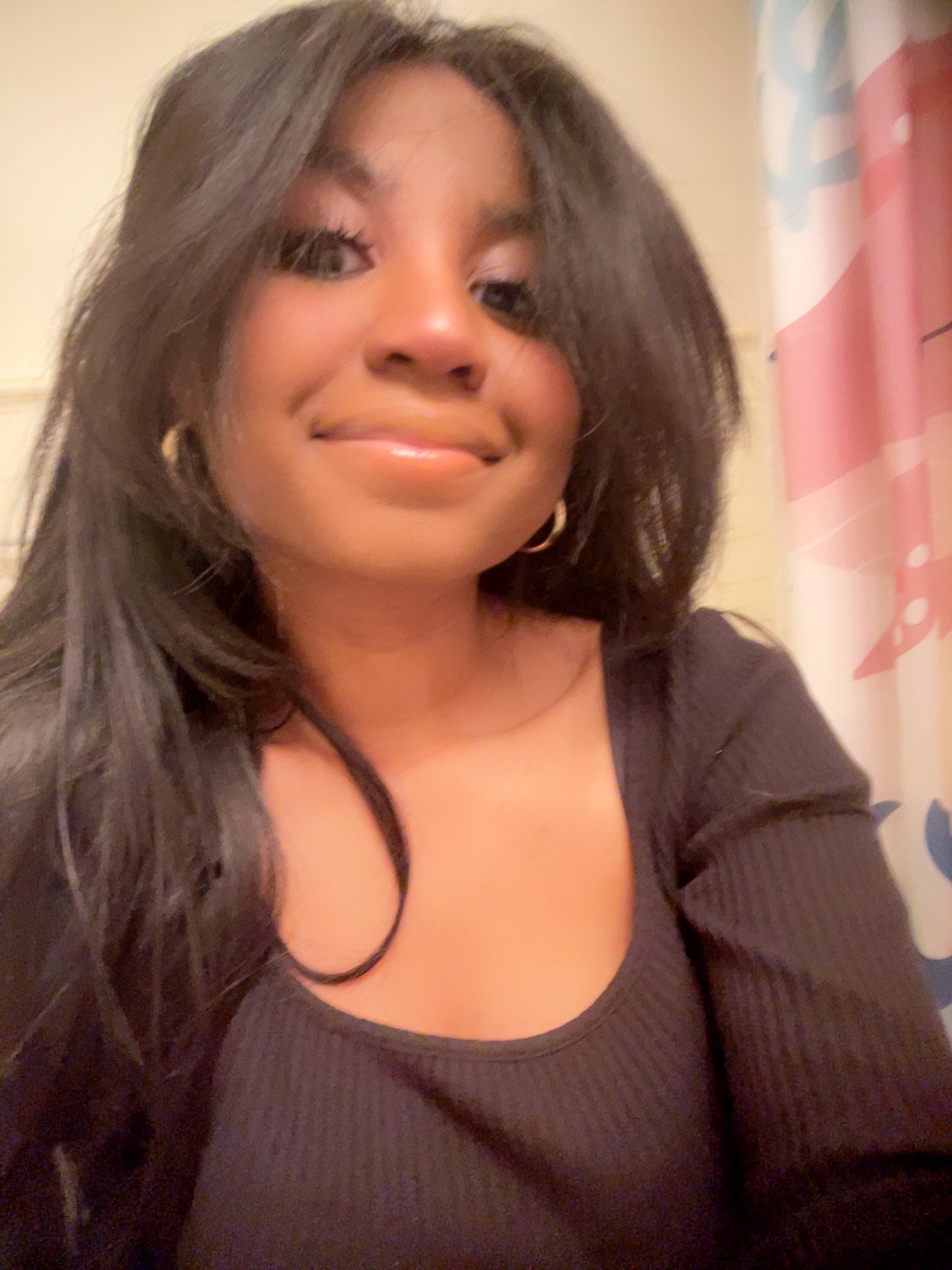 Close-up of a young woman with long dark hair, wearing gold hoop earrings and a black top, smiling at the camera.