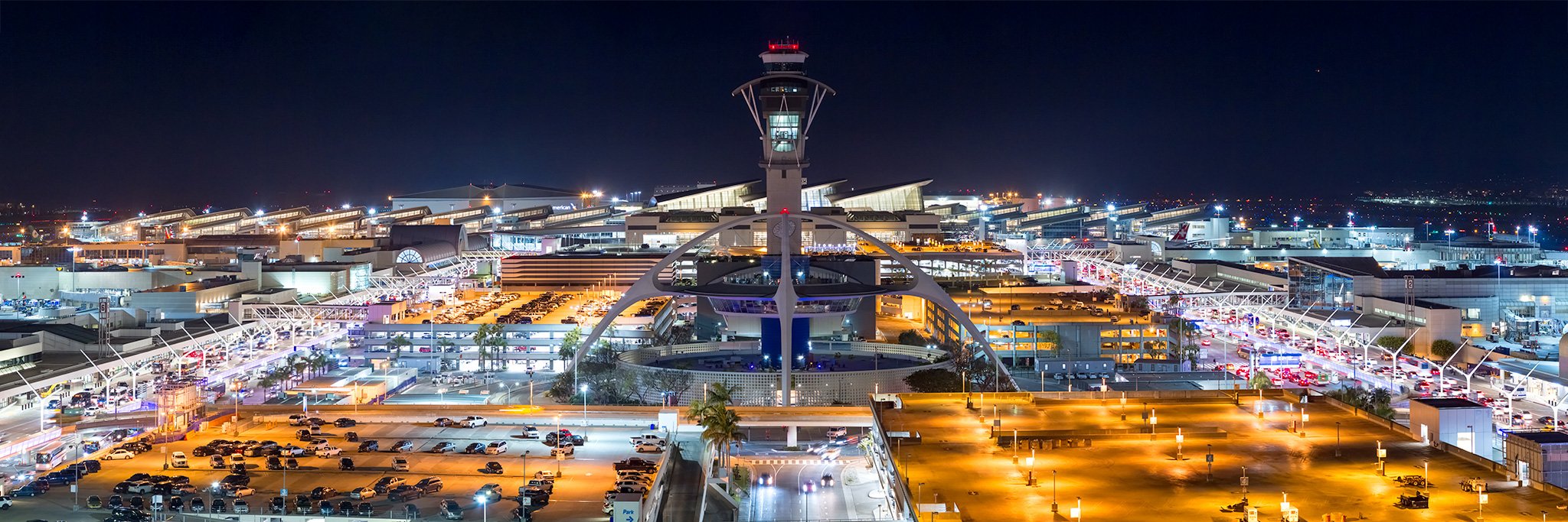 Aerial view of a large airport terminal at night with a central control tower, illuminated parking lots, and surrounding structures. Bright lights and car traffic are visible.