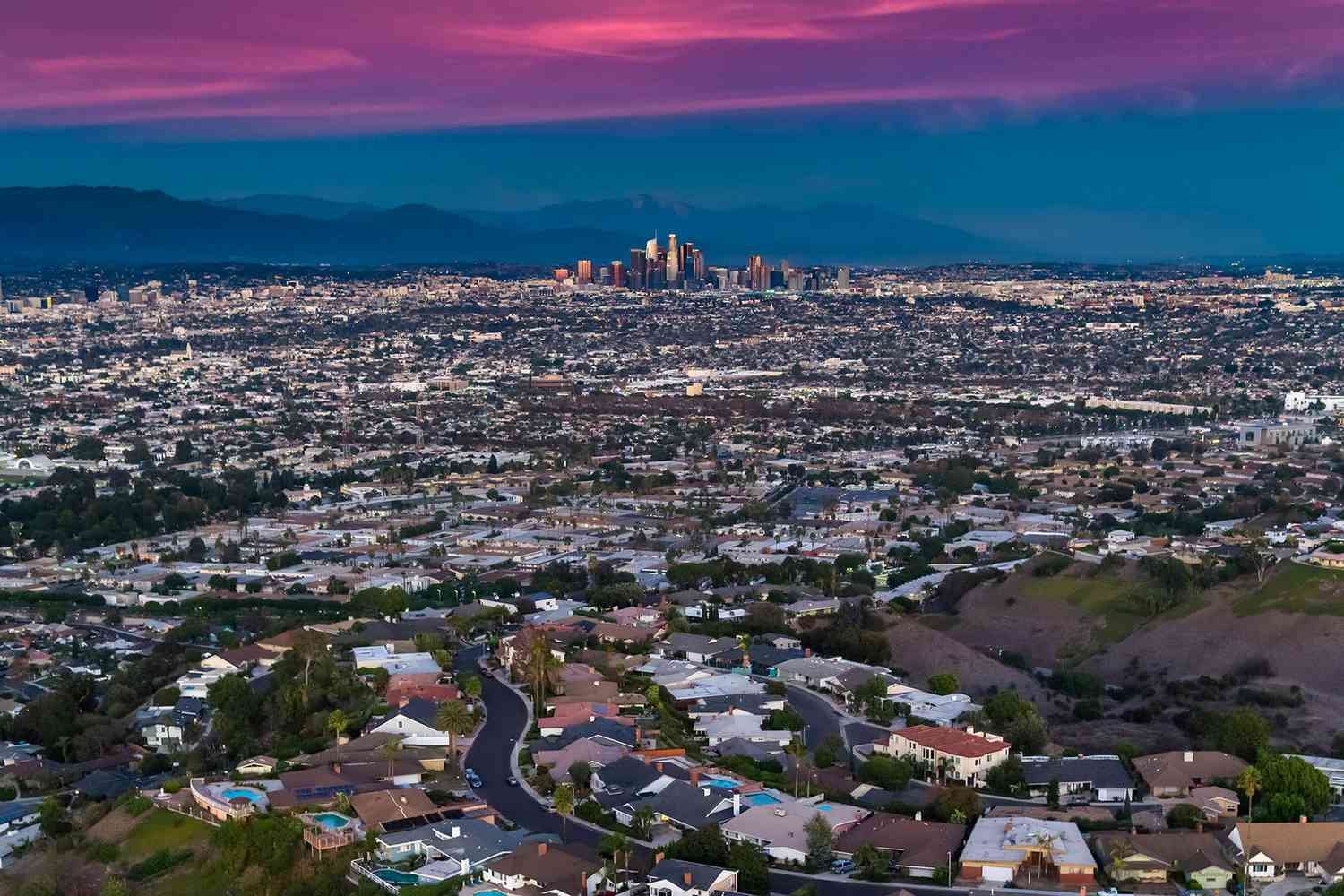 Aerial view of Los Angeles cityscape at sunset with residential neighborhood in foreground and downtown skyline in background.