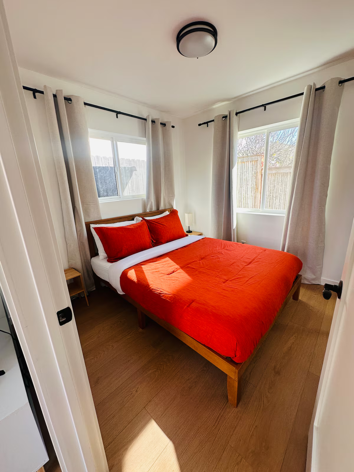 Small bedroom with a wooden bed frame, red bedding, white walls, and two windows with beige curtains.