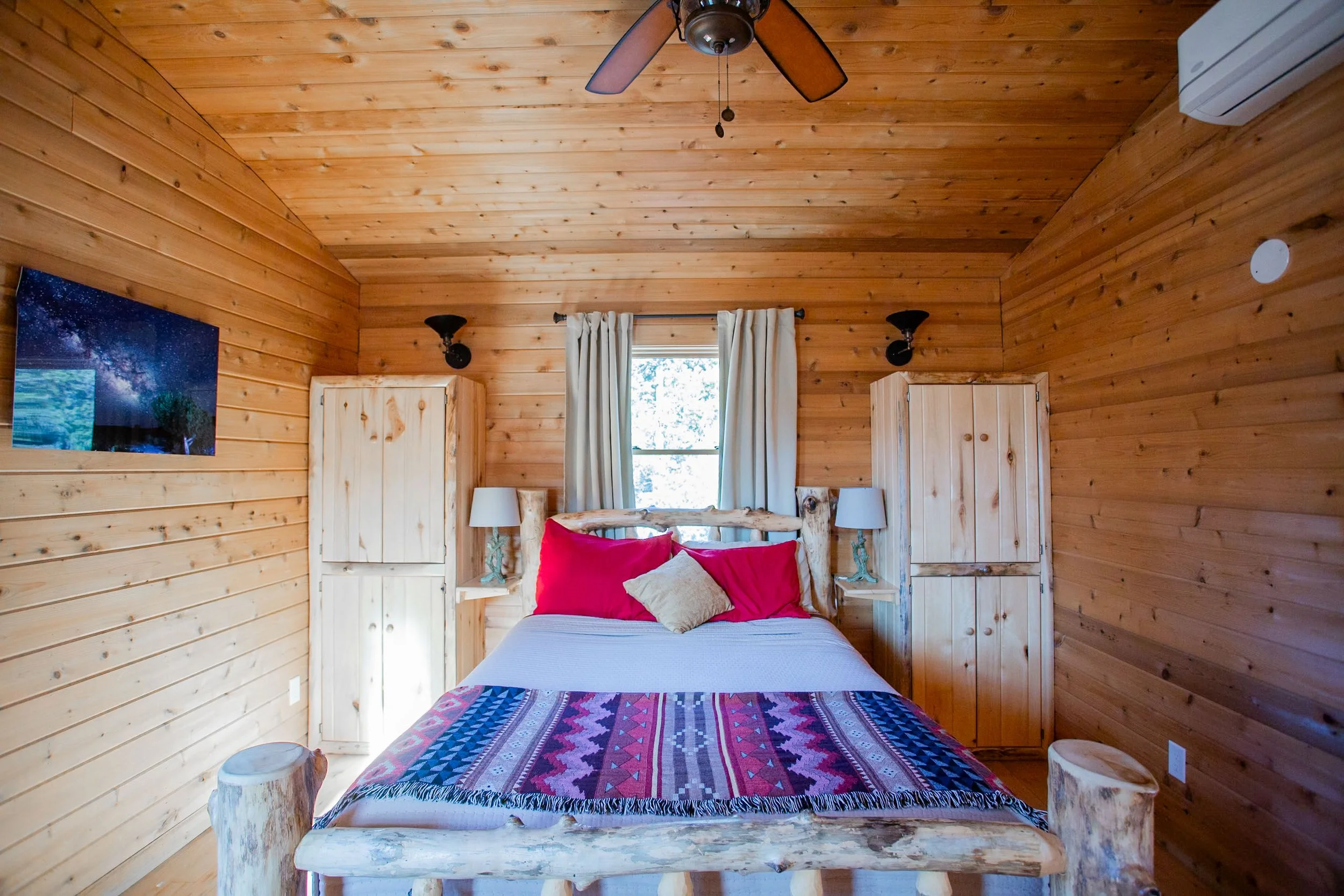 Interior of a rustic bedroom with wood-paneled walls and ceiling, featuring a bed with colorful patterned bedding and red pillows, two nightstands with white lamps, a window with white curtains, wall-mounted black lamps, a ceiling fan, and part of a wall-mounted artwork depicting a starry night sky.