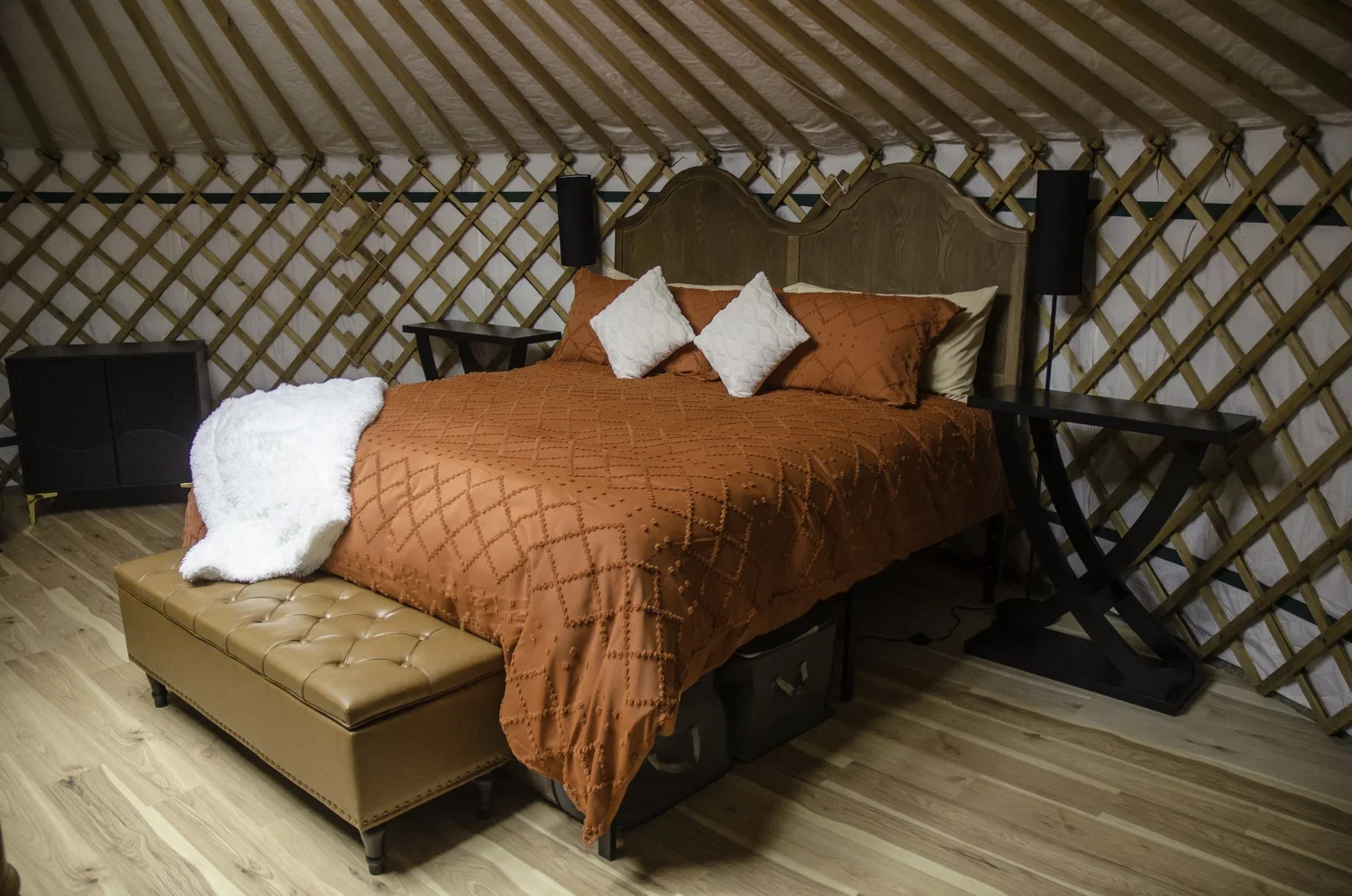 A cozy bedroom with a wooden bed frame, orange quilted bedding, two white pillows, a tan leather bench, and wooden floor, with decor yurt-style walls.