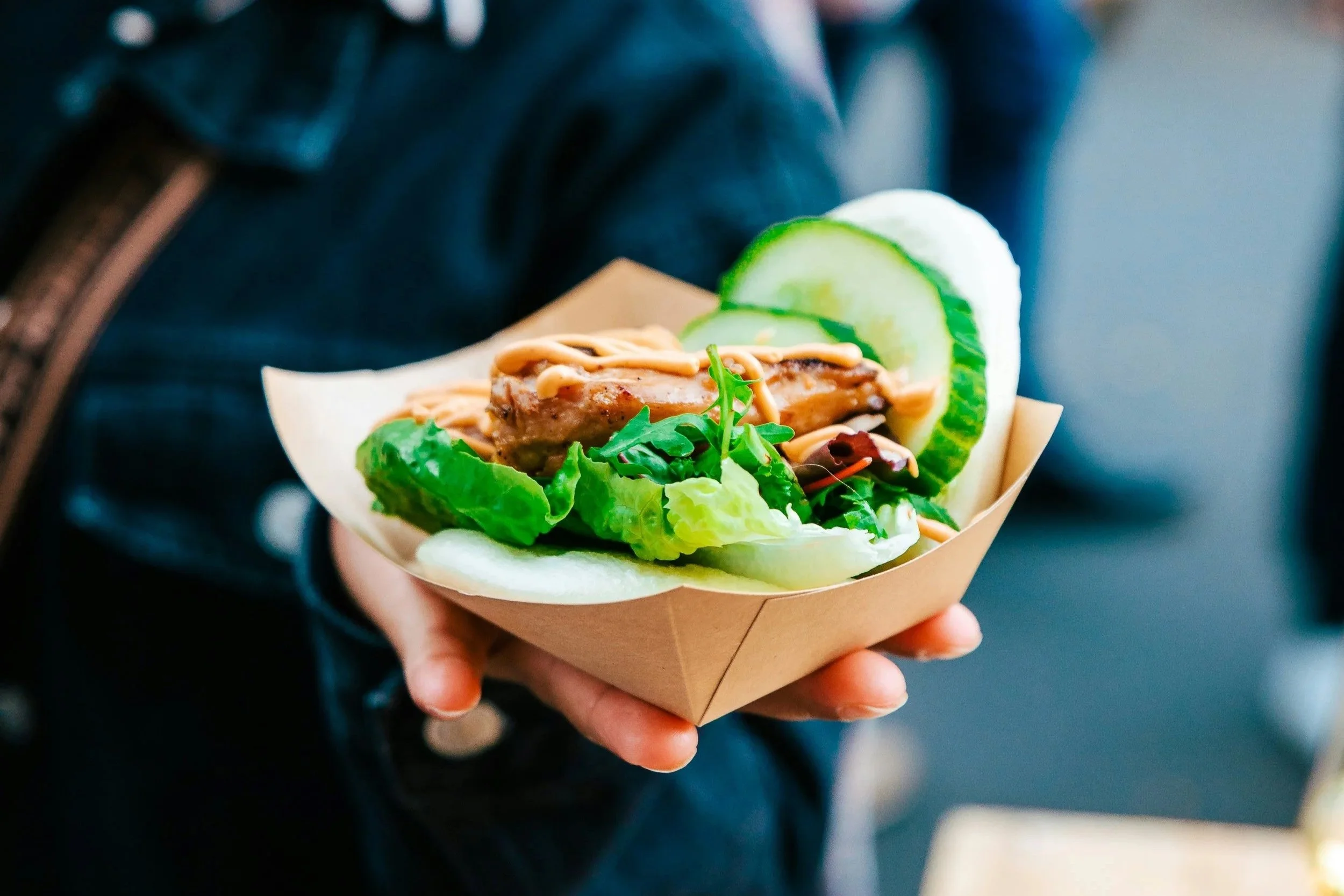 A person holding a paper food tray with a cucumber and salad wrap, lettuce, and some cucumber slices.