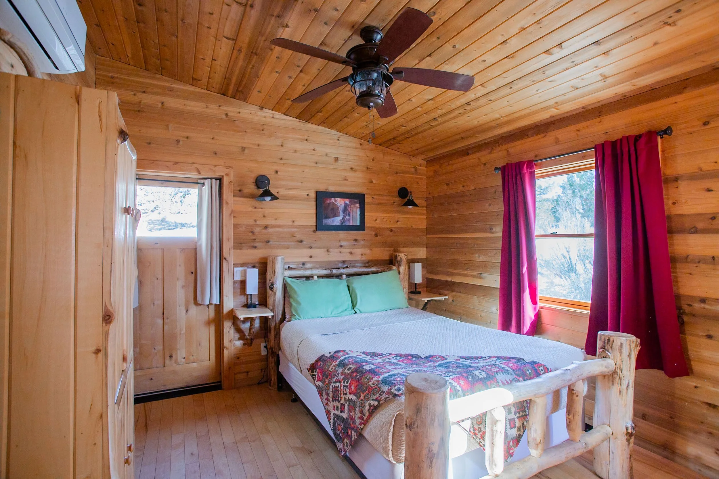 A rustic bedroom with wood-paneled walls and ceiling, a log bed with green pillows, large windows with red curtains, a ceiling fan, and framed art on the wall.