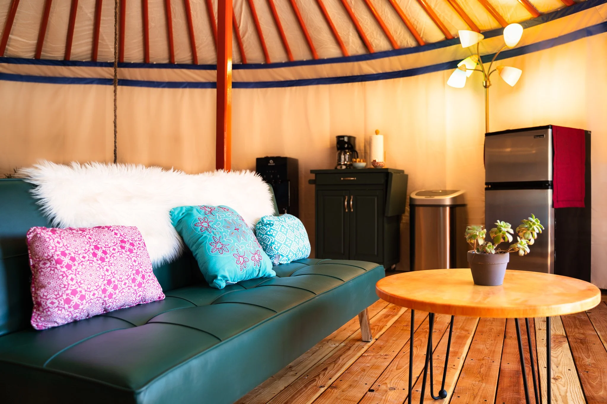 A cozy living space with a green couch decorated with colorful and patterned pillows, a round wooden coffee table, a small green cabinet, a mini refrigerator, a coffee maker, and a floor lamp with multiple white lampshades against a curved wooden ceiling and wooden floor.