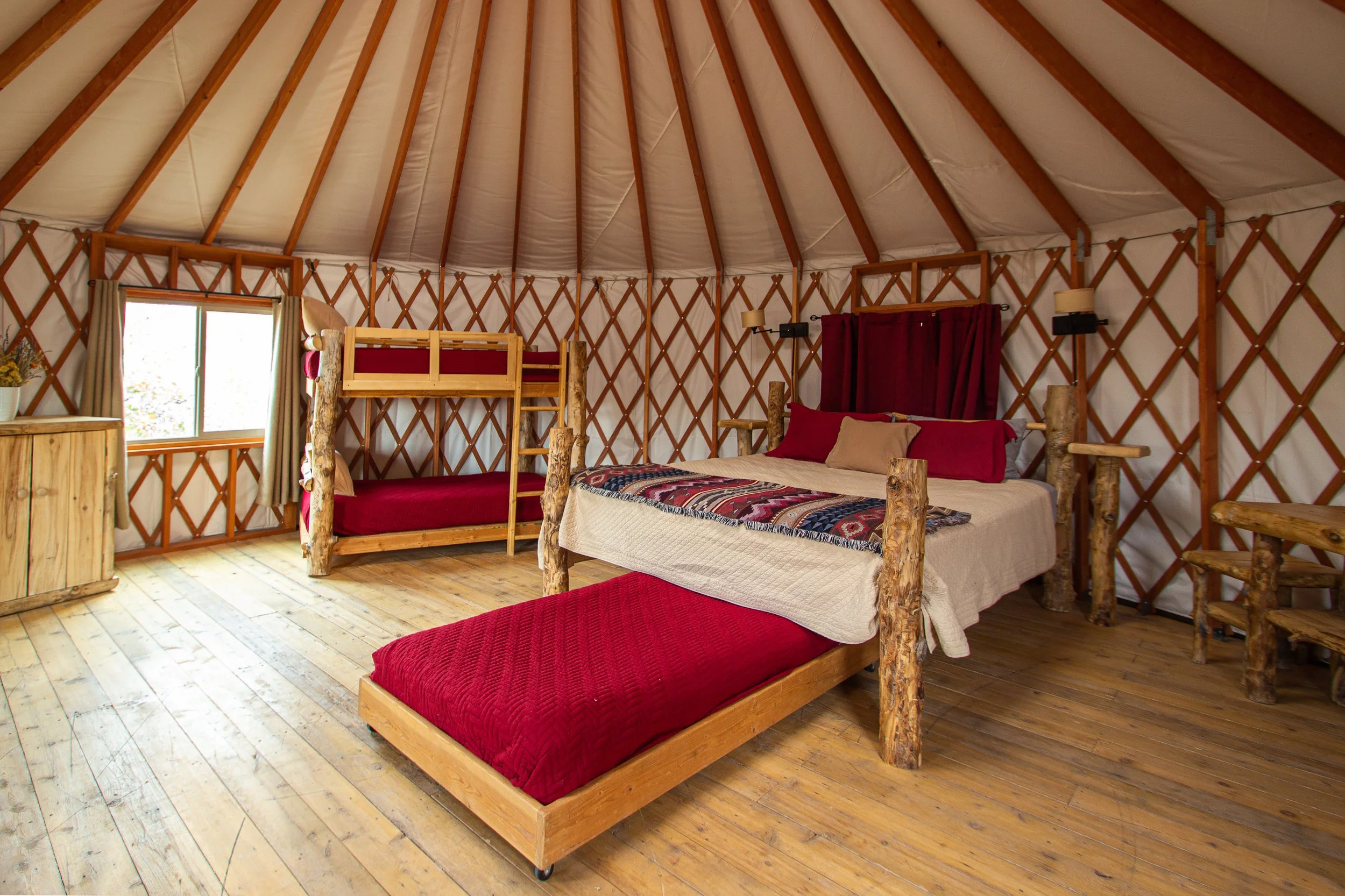 Interior of a yurt with a wooden bed, a bunk bed, and red and beige bedding, wood flooring, and windows with curtains.